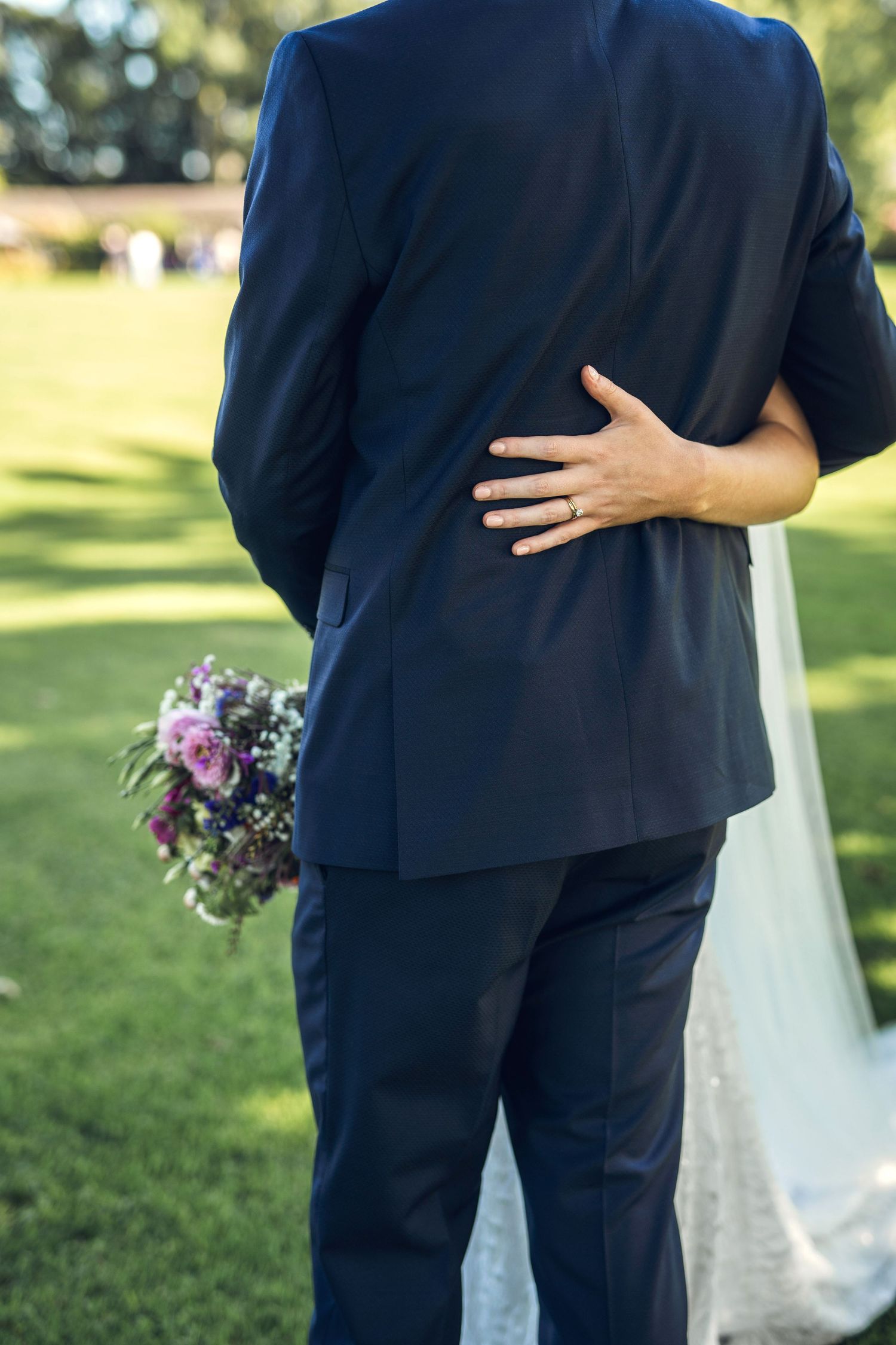 Close-up of bride placing ringed hand on grooms navy suit back while holding purple wedding bouquet outdoors.