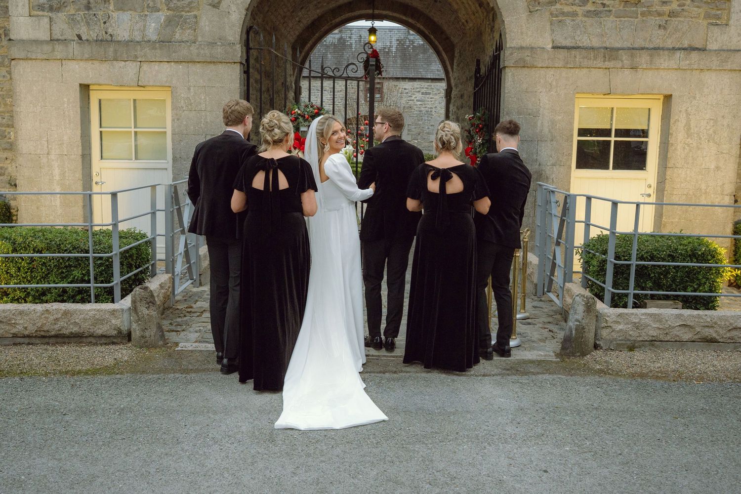 Wedding party in black velvet attire stands with bride before stone archway entrance at venue.