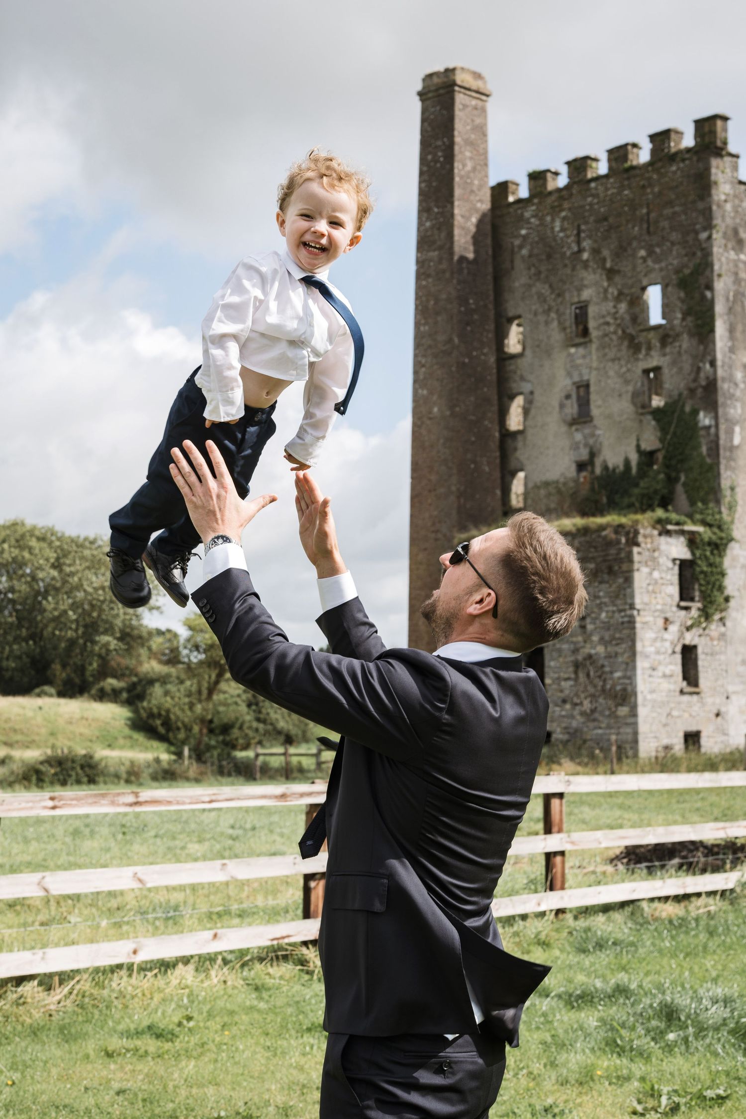 Groom in dark suit lifting a laughing young boy in the air outside a historic castle.