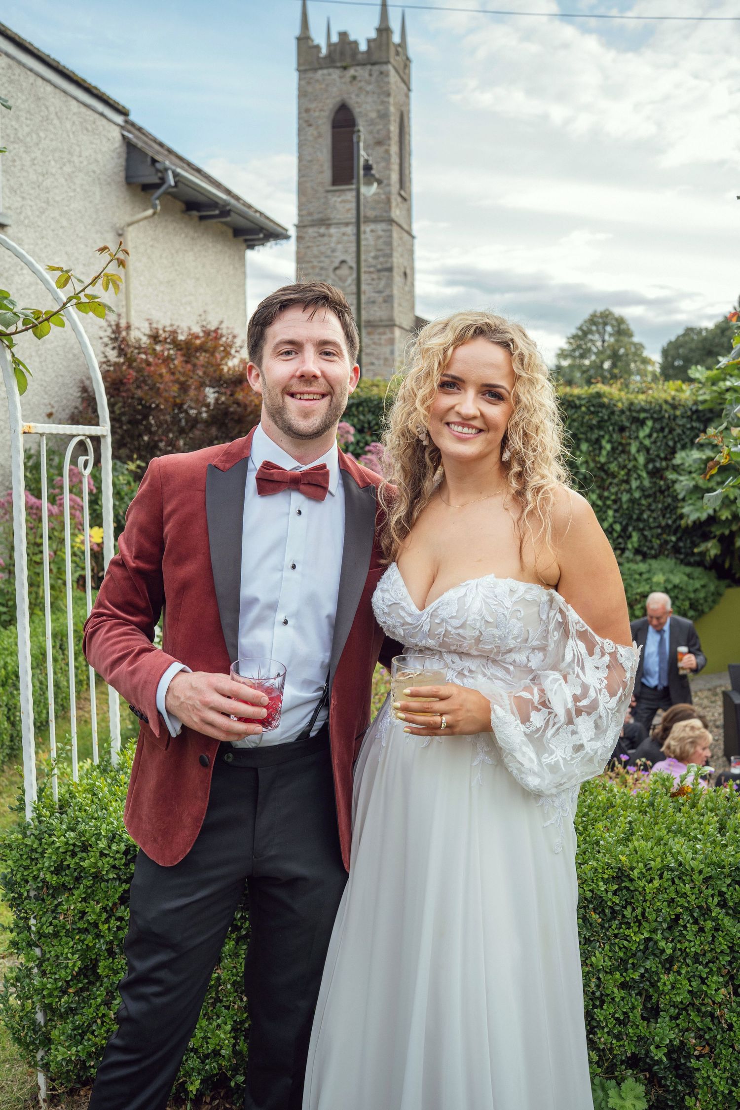 Smiling bride in white lace off-shoulder gown and groom in burgundy tuxedo pose at outdoor wedding at garden wedding reception in Co Meath.