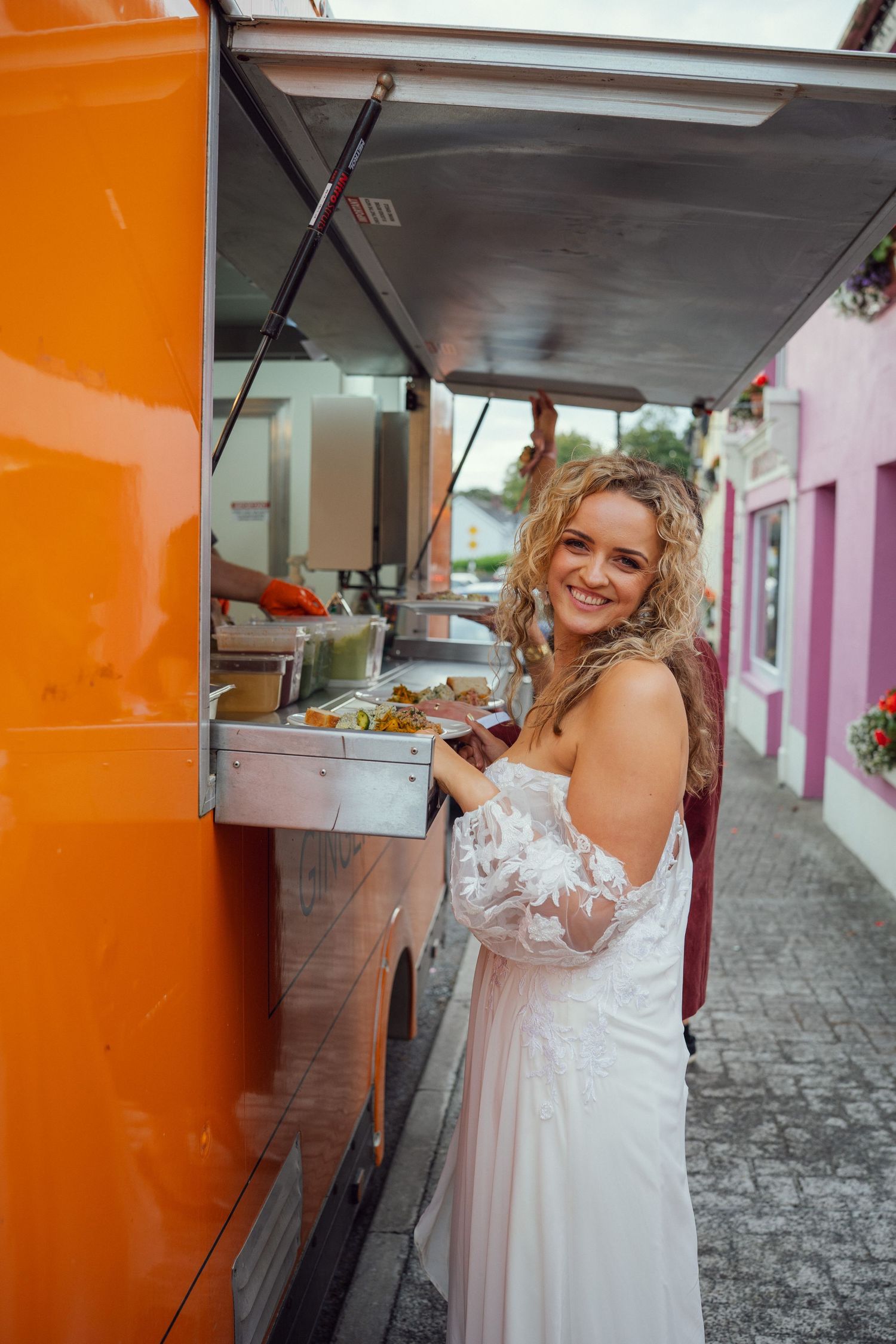 A smiling bride in a white lace dress ordering food from a bright orange food truck at outdoor garden wedding reception in Co Meath.