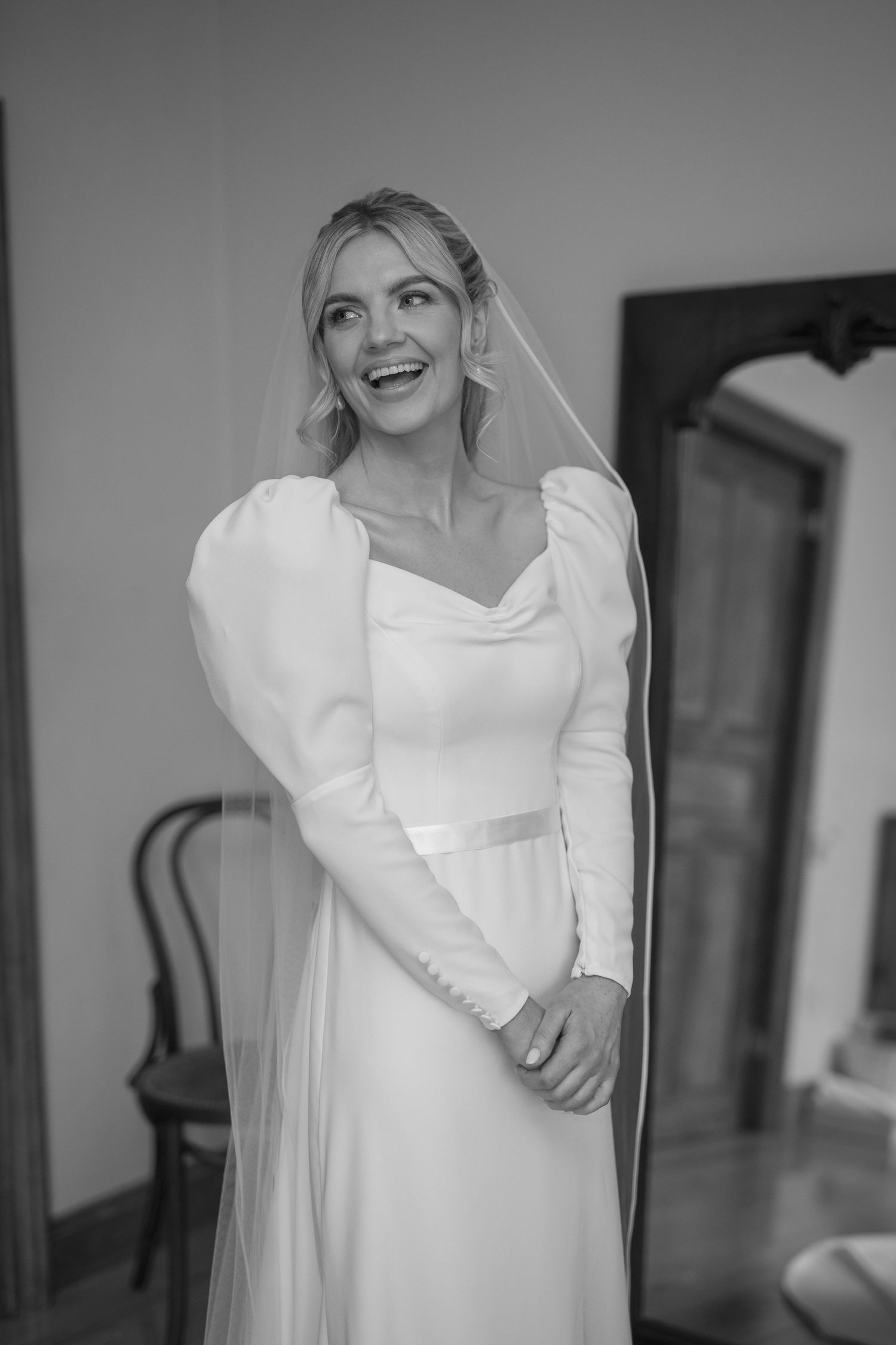 Radiant bride in long-sleeve white gown and veil laughs in black and white portrait.