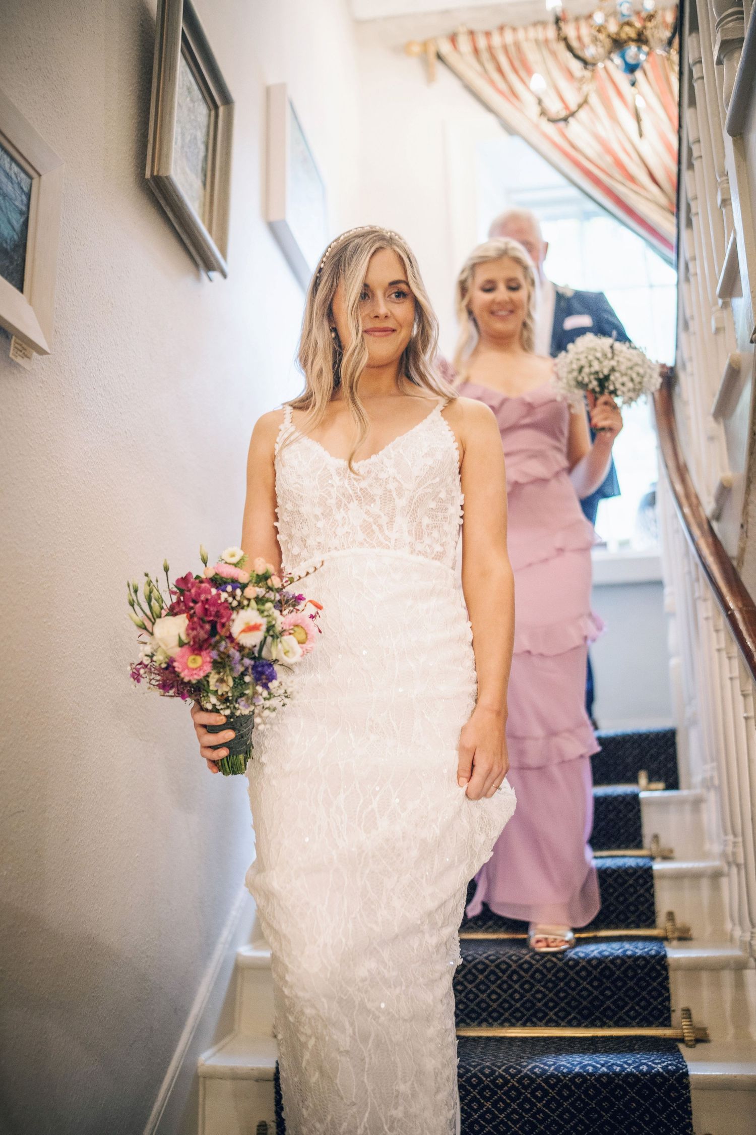 Elegant bride in lace gown descending staircase with bridesmaid and guest behind her.