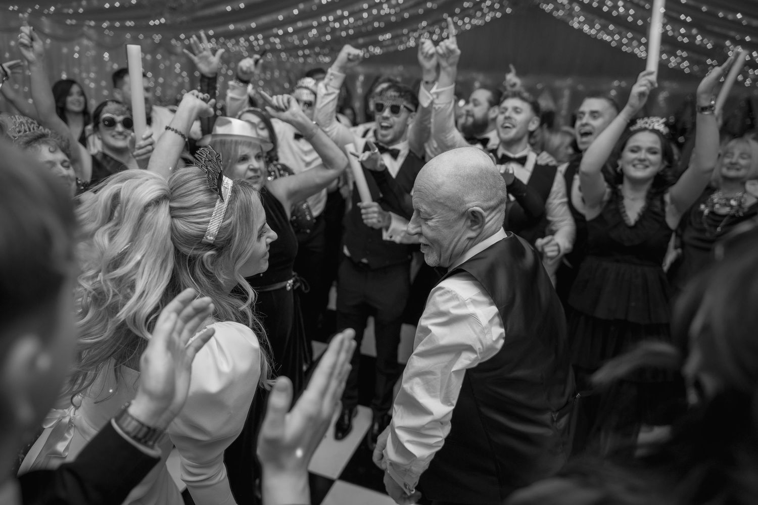 Black and white photo of bride and bald man dancing energetically at crowded wedding reception.