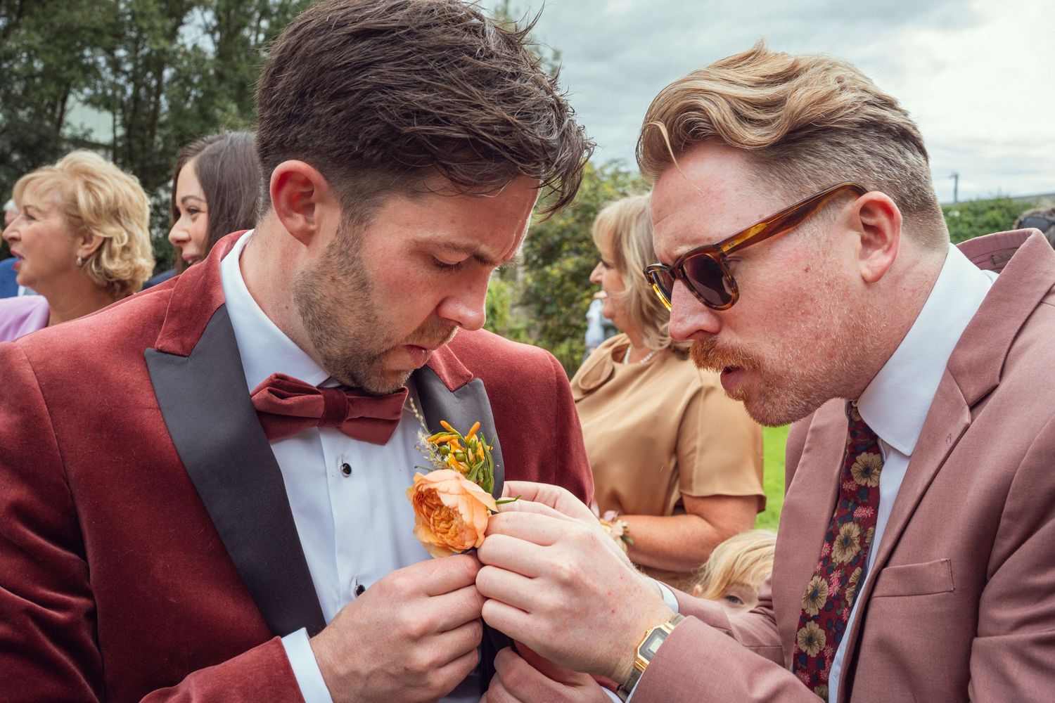 Groom in burgundy tuxedo has boutonniere pinned by groomsman at outdoor garden wedding reception in Co Meath.