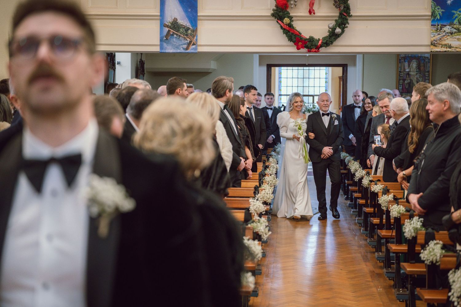 Bride walks down church aisle with escort past rows of standing wedding guests in formal attire.