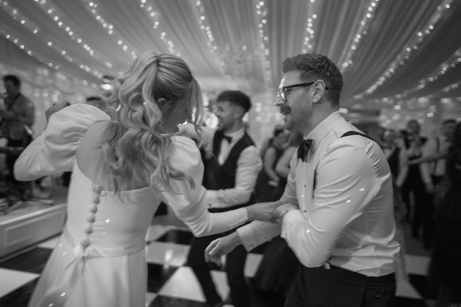 Bride and groom share a joyful first dance in black and white wedding photo under twinkling lights.