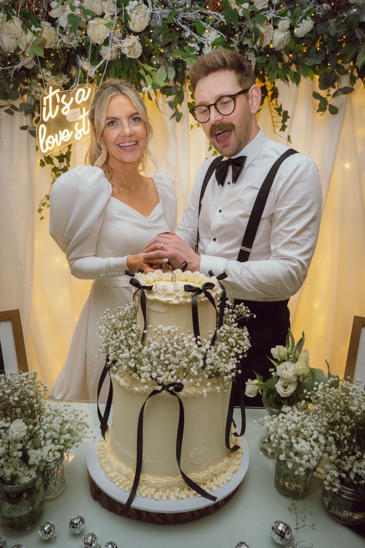Smiling bride and groom cutting their tiered white wedding cake beneath a floral arch.