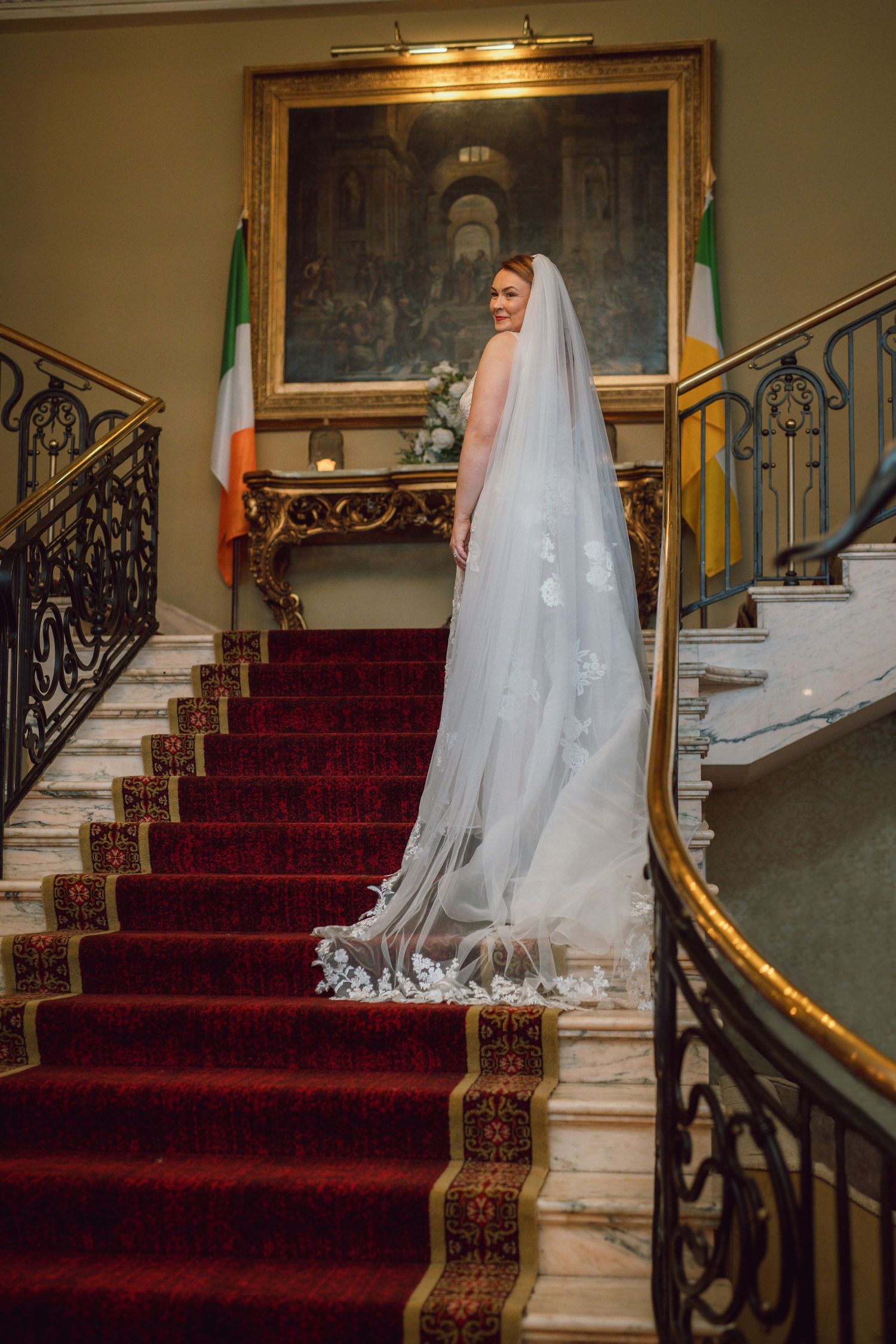 Bride in lace wedding gown with long veil standing on red carpeted grand staircase.