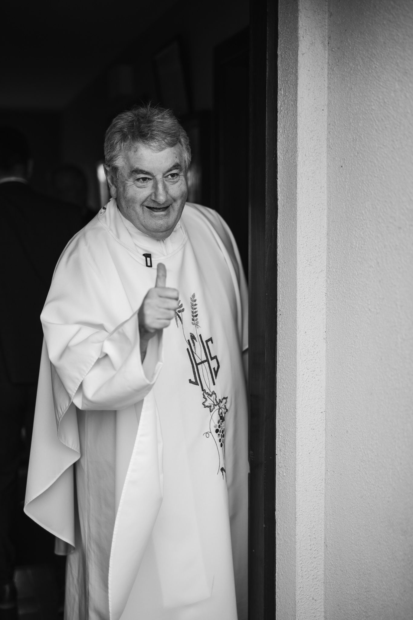 Black and white photo of smiling priest in white vestments giving thumbs up at church doorway.