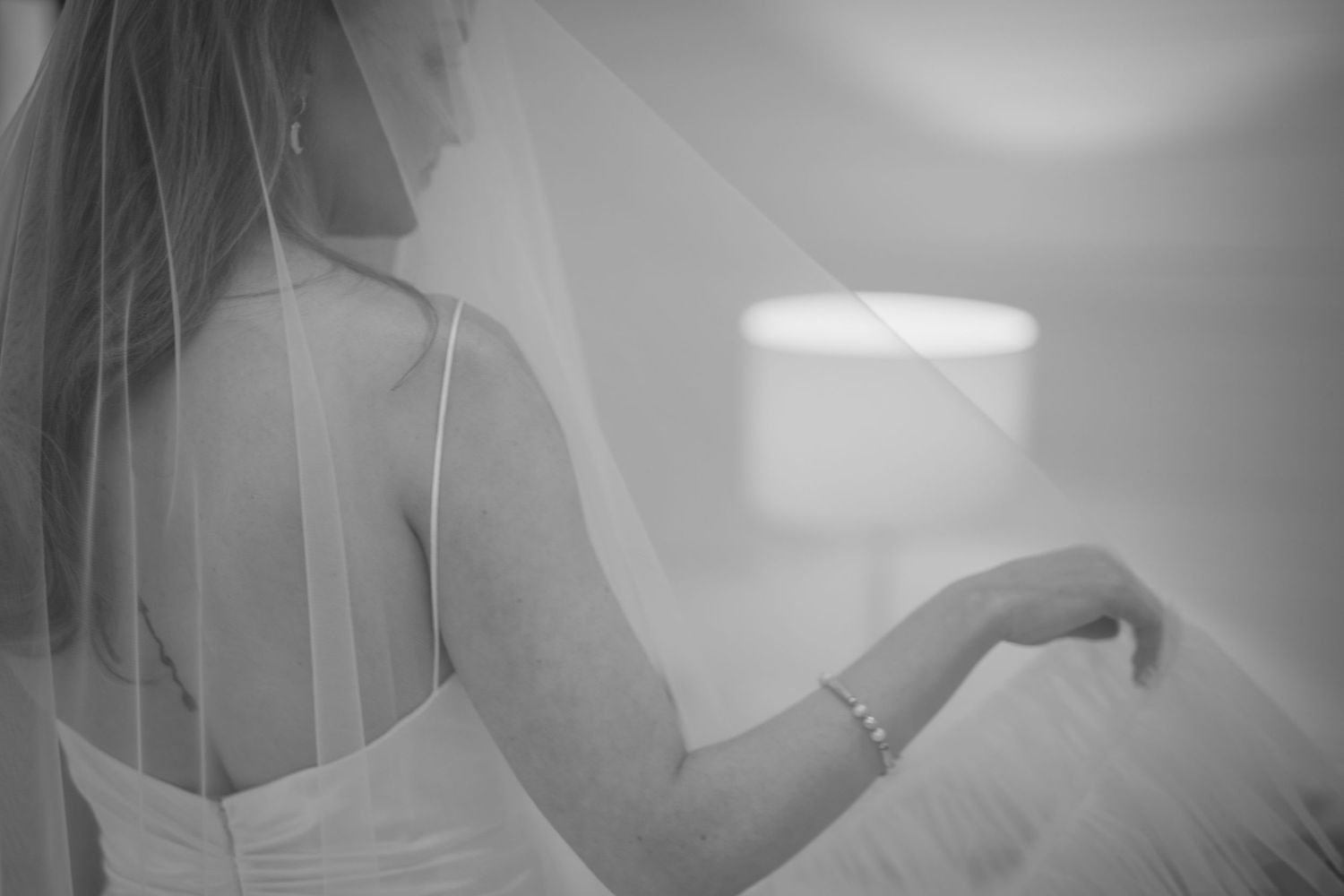 Black and white photo of bride in wedding dress and veil viewed from behind in soft natural light.