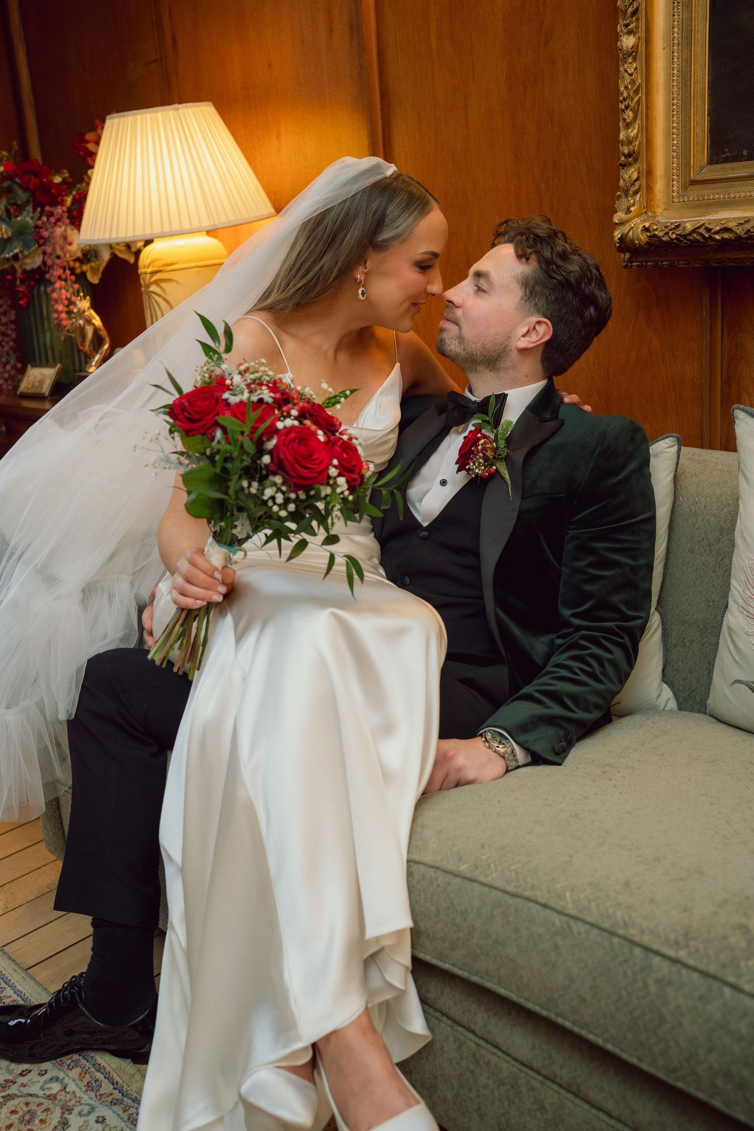 Bride and groom sharing romantic moment on sofa with red bouquet.