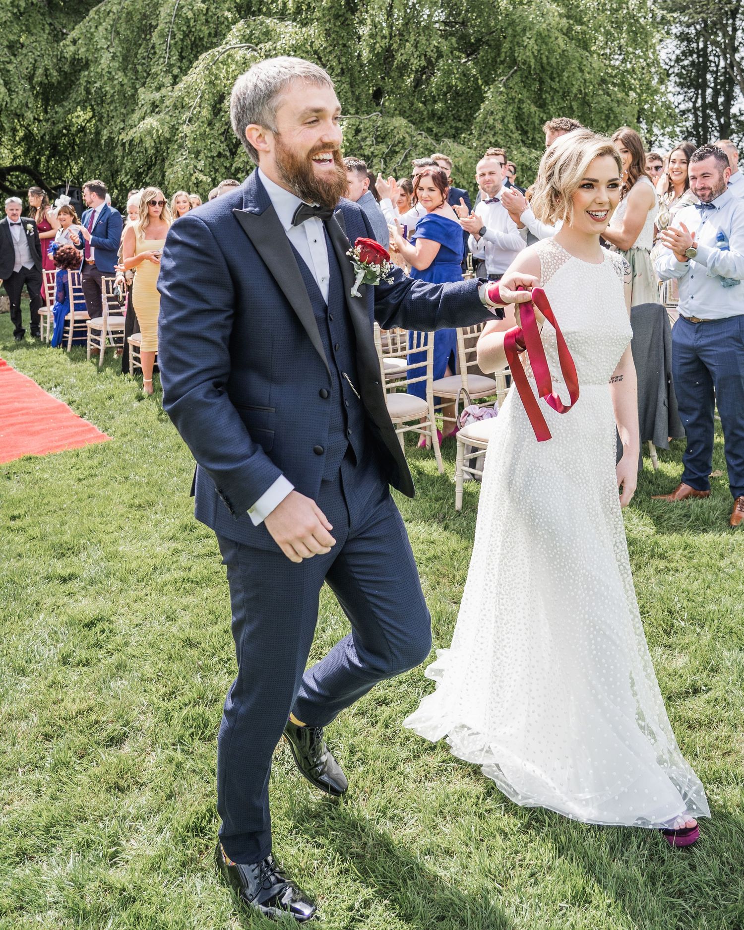 Newlyweds walk triumphantly down grass aisle, groom in navy suit, bride in lace gown holding red ribbon.