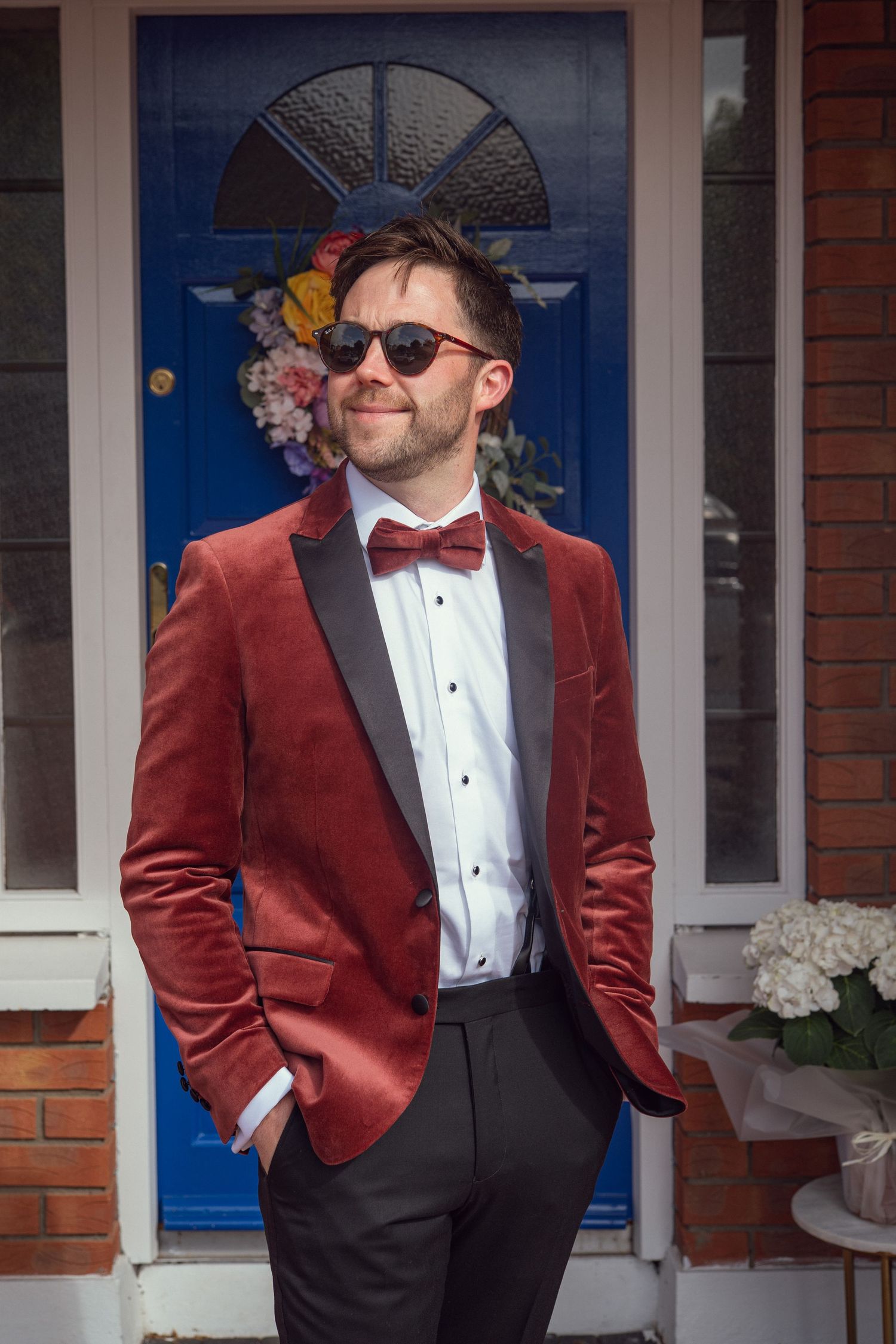 Stylish groom in red velvet tuxedo and sunglasses poses confidently in front of blue front door.
