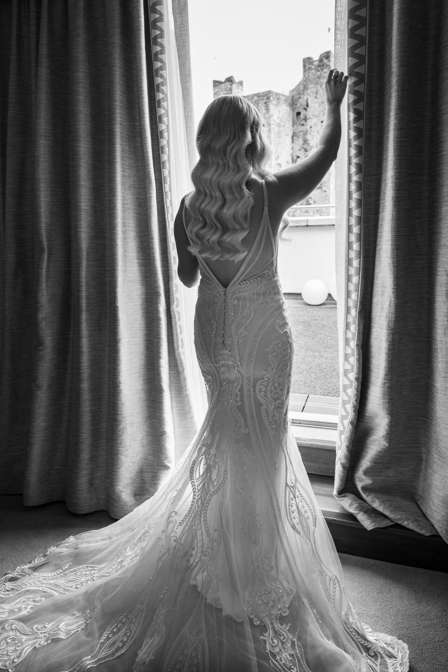 Elegant bride in fitted lace mermaid gown with train stands gazing out window in black and white.