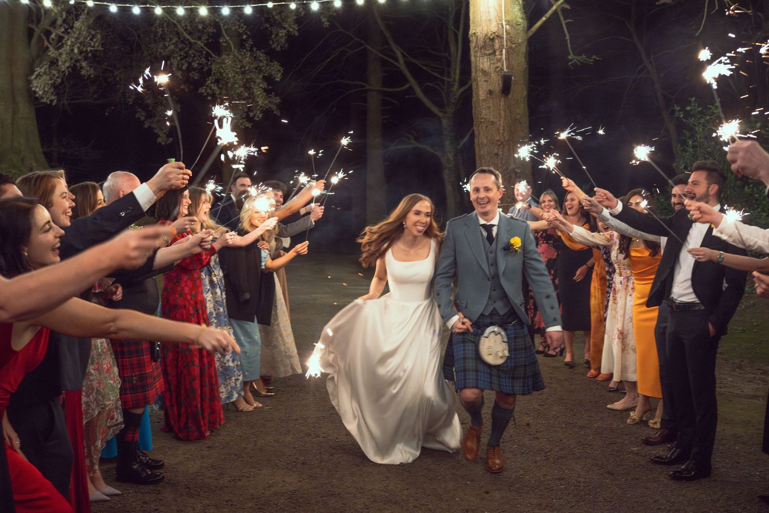 Bride and groom in Scottish kilt walking through a sparkler tunnel surrounded by cheering wedding guests at night.