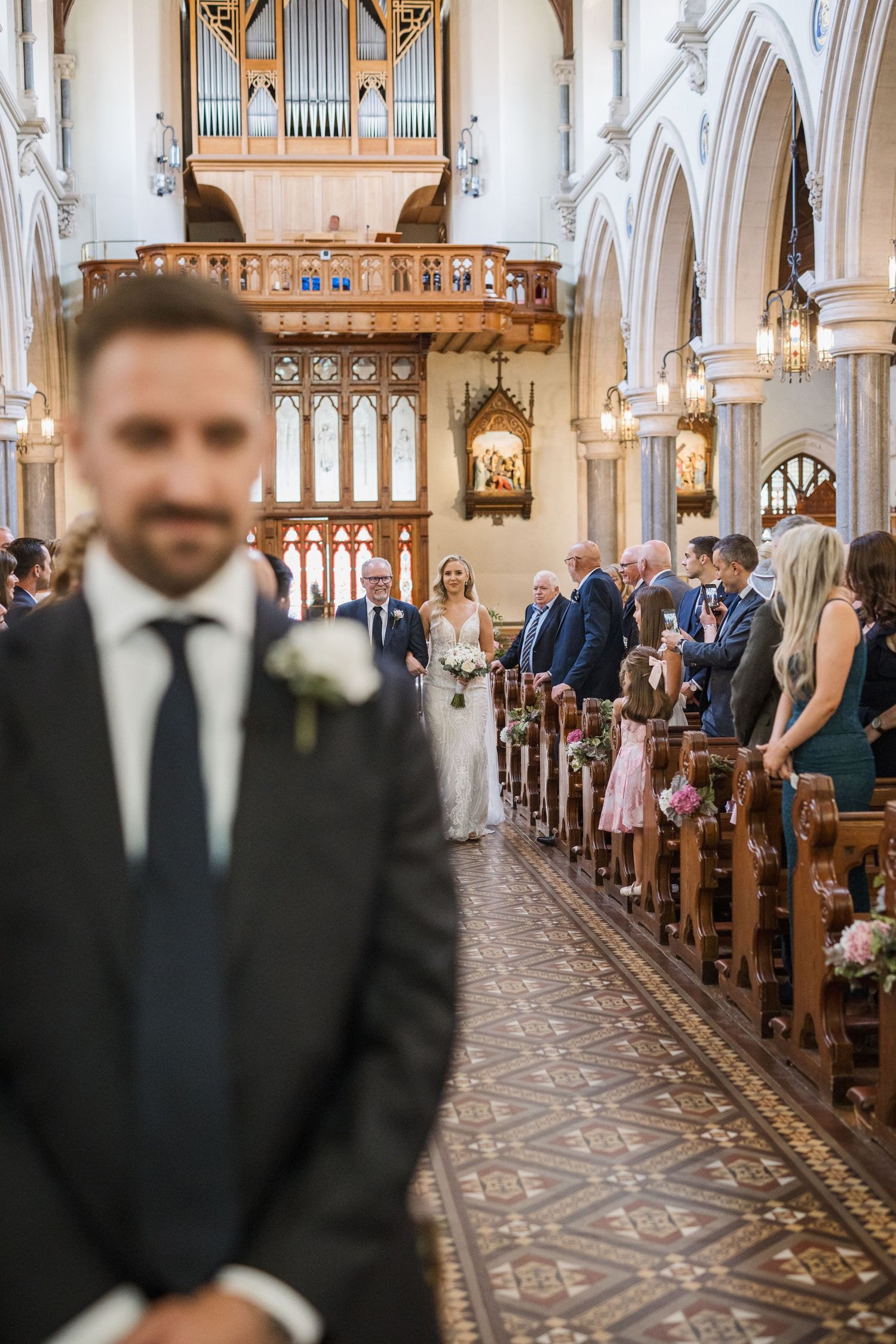 Groom stands in foreground as bride walks down the aisle of a grand church during wedding ceremony.