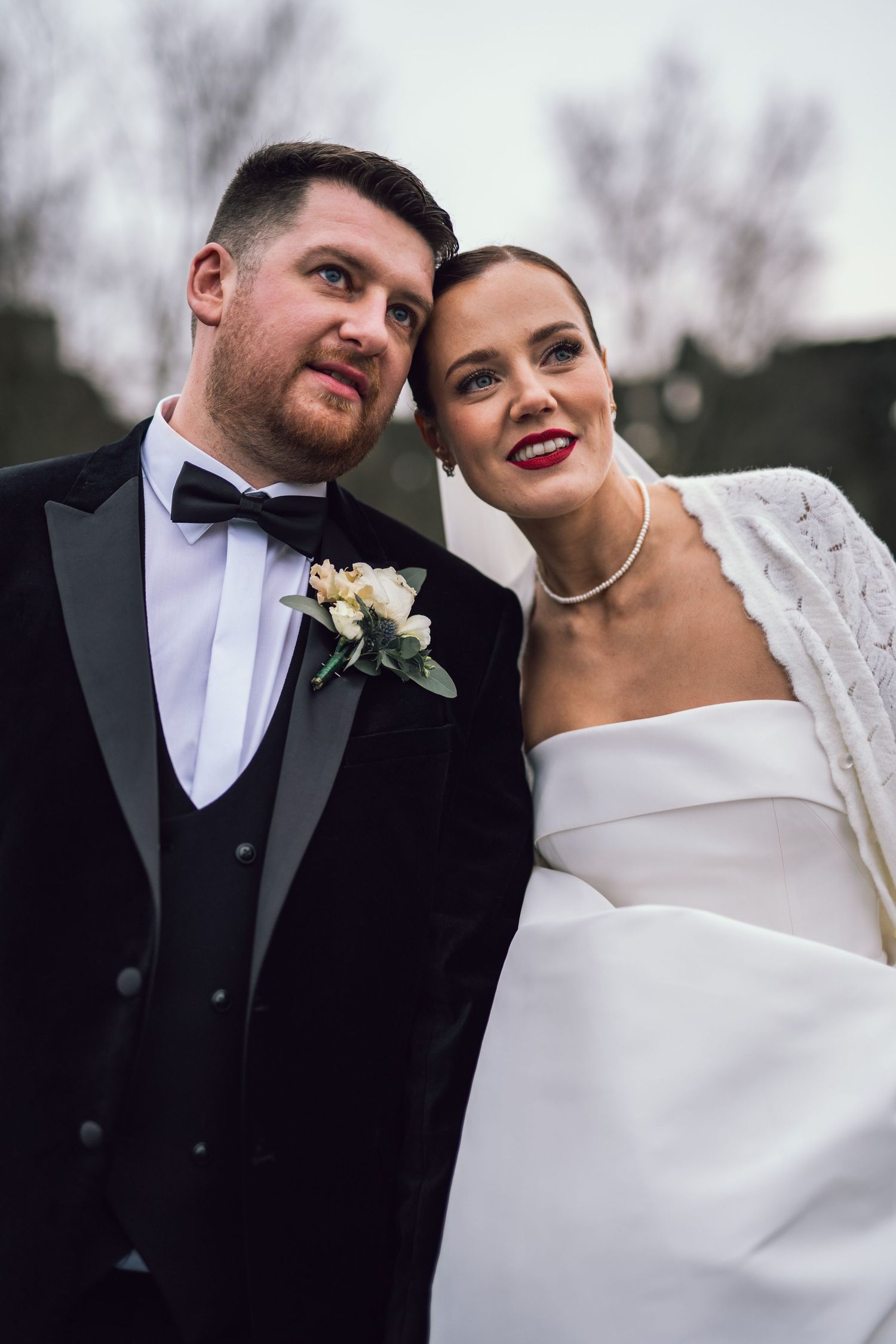 Bride in white strapless gown with pearl necklace and groom in black tuxedo pose outdoors.