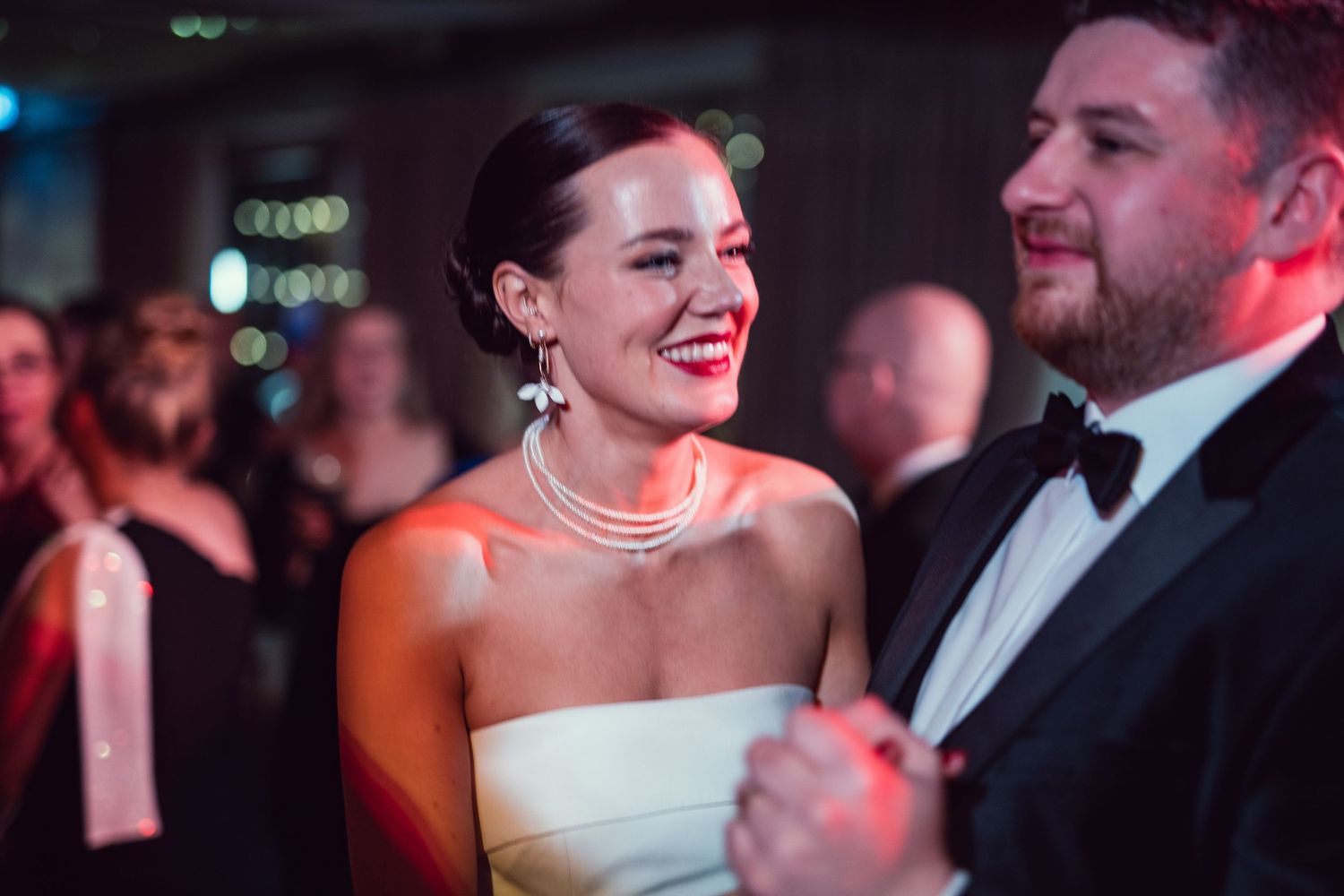 Bride in strapless white gown and pearl necklace laughing joyfully with groom in tuxedo at elegant wedding reception.