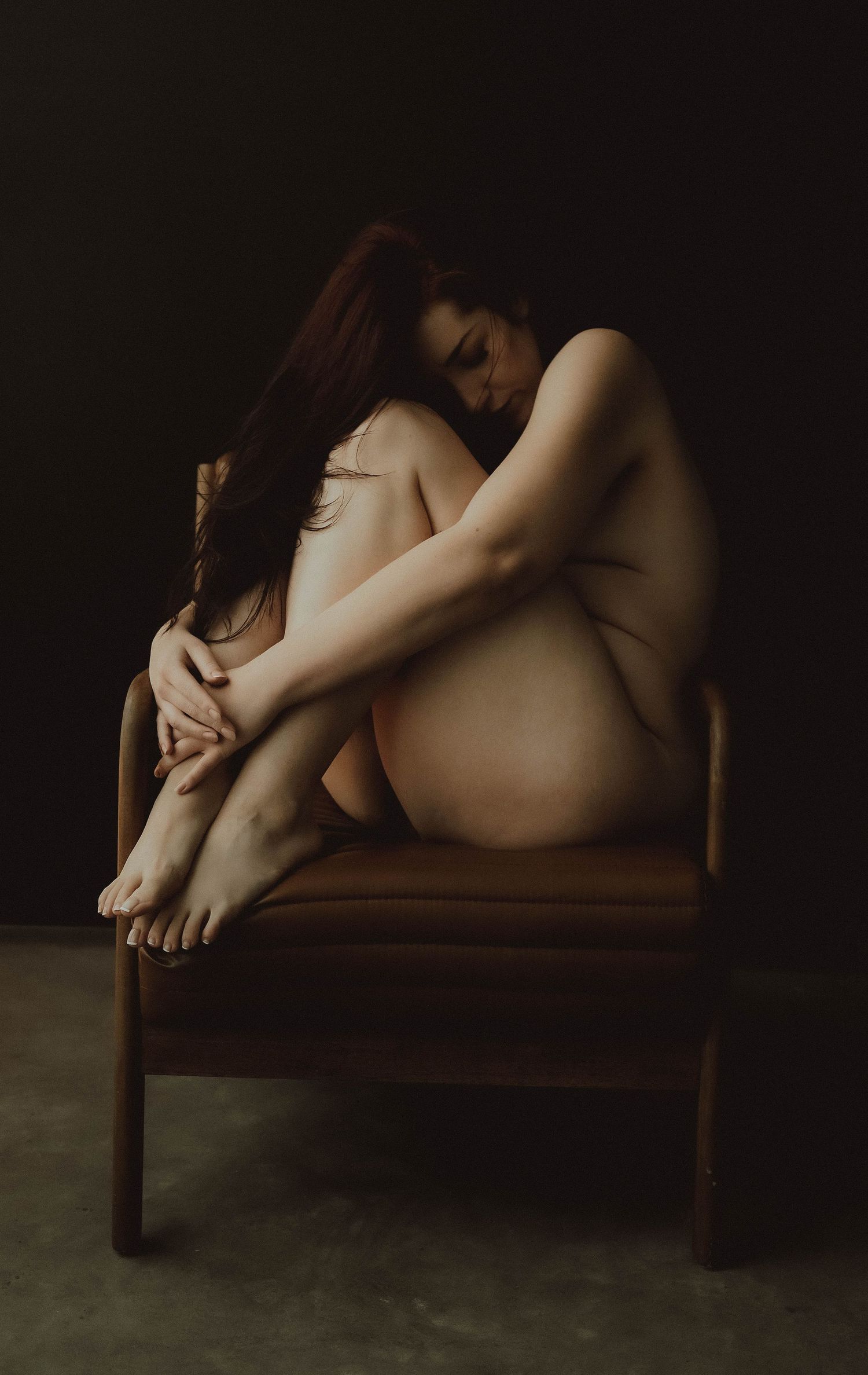 Artistic nude figure study showing graceful curves and shadows on a vintage wooden chair in moody lighting.
