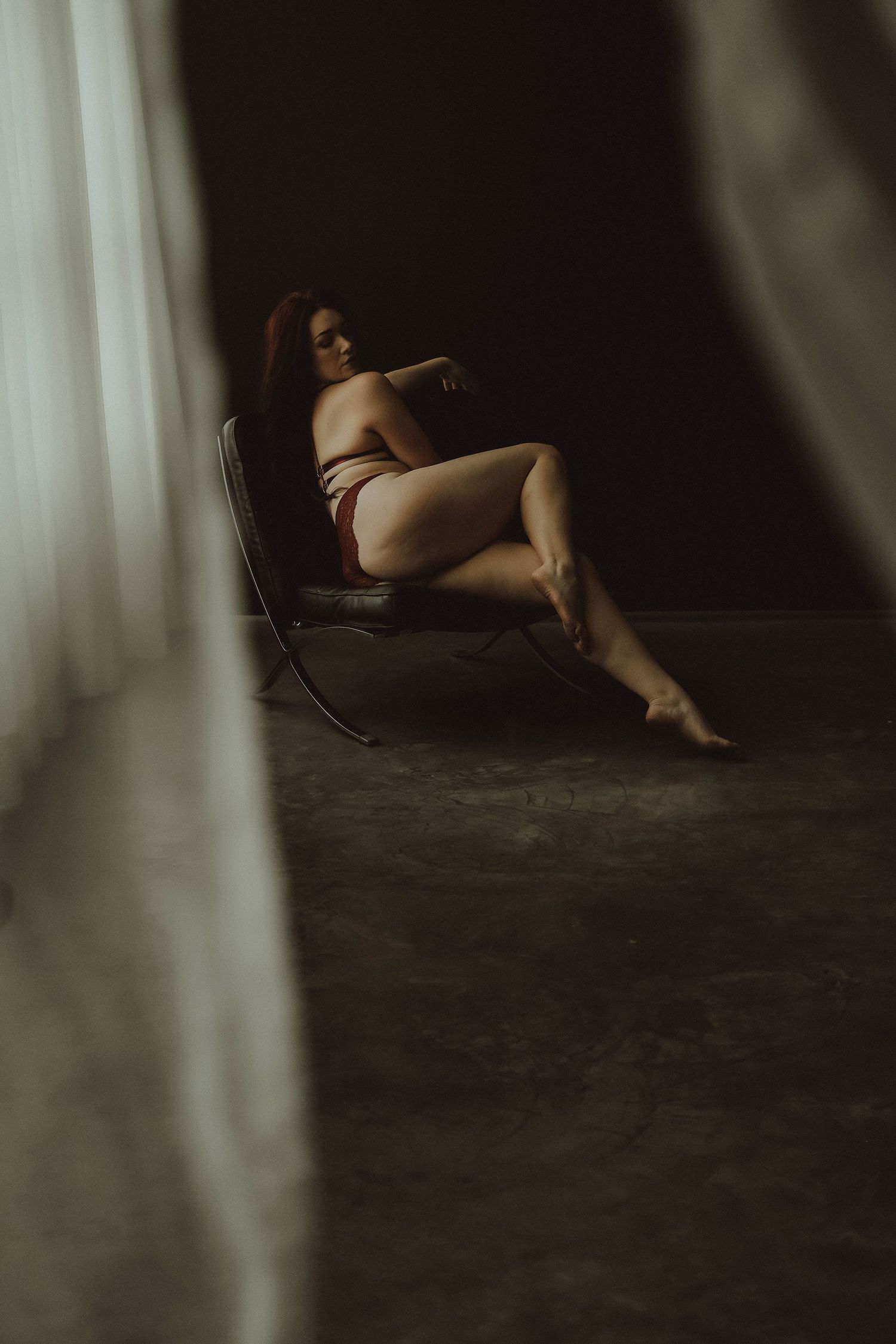 Moody boudoir photograph of a person reclining in lingerie on a chair with dramatic window lighting and shadows.