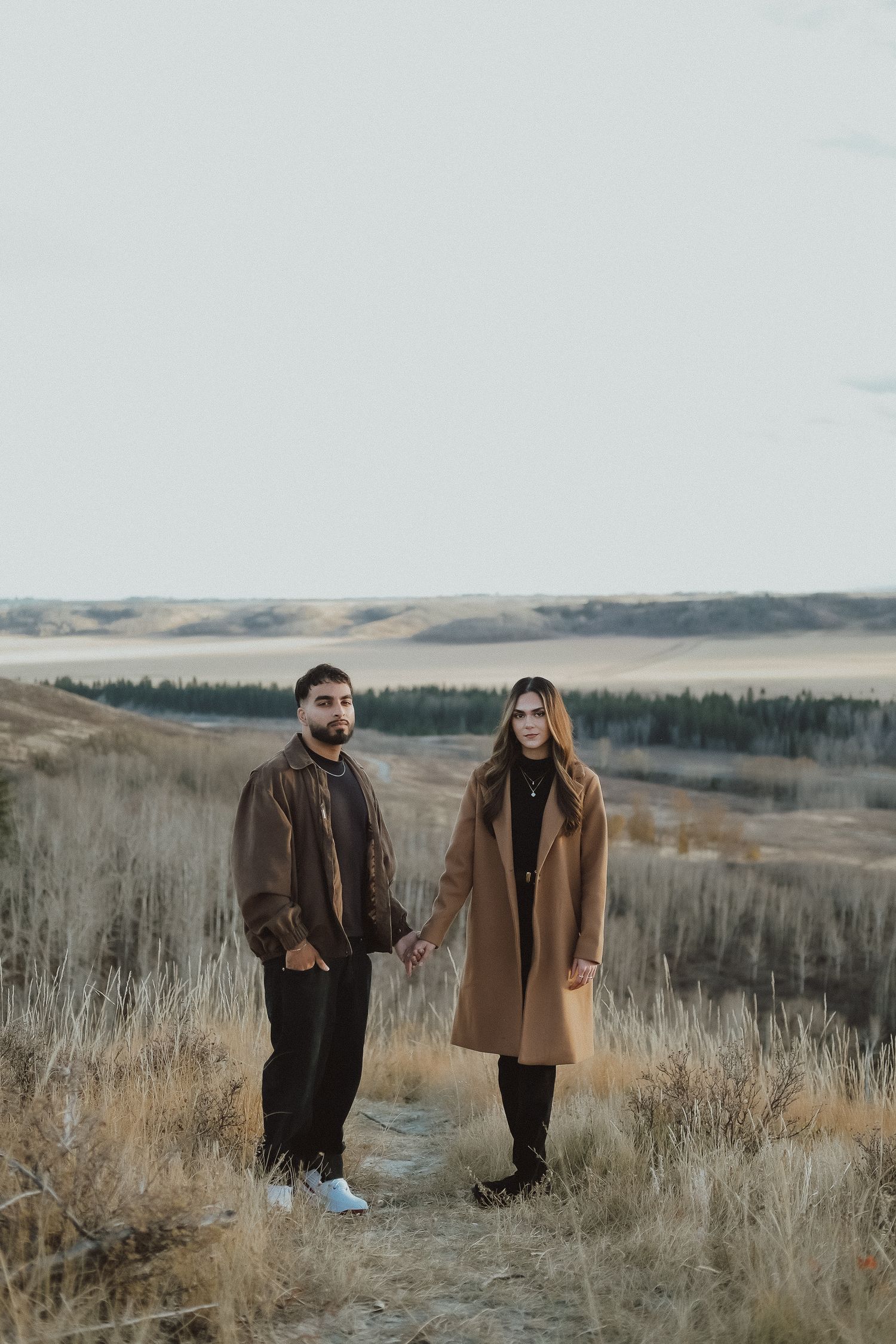 Two people in brown coats walk through a serene autumn field with tall grass and distant hills at sunset.