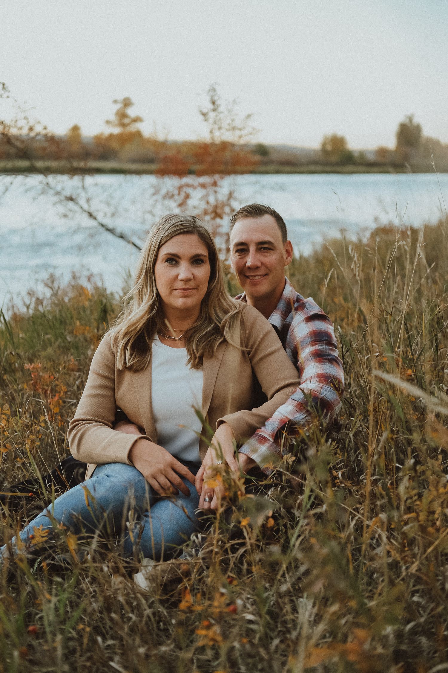 A couple sits together in tall grass by a lake during golden hour in autumn.