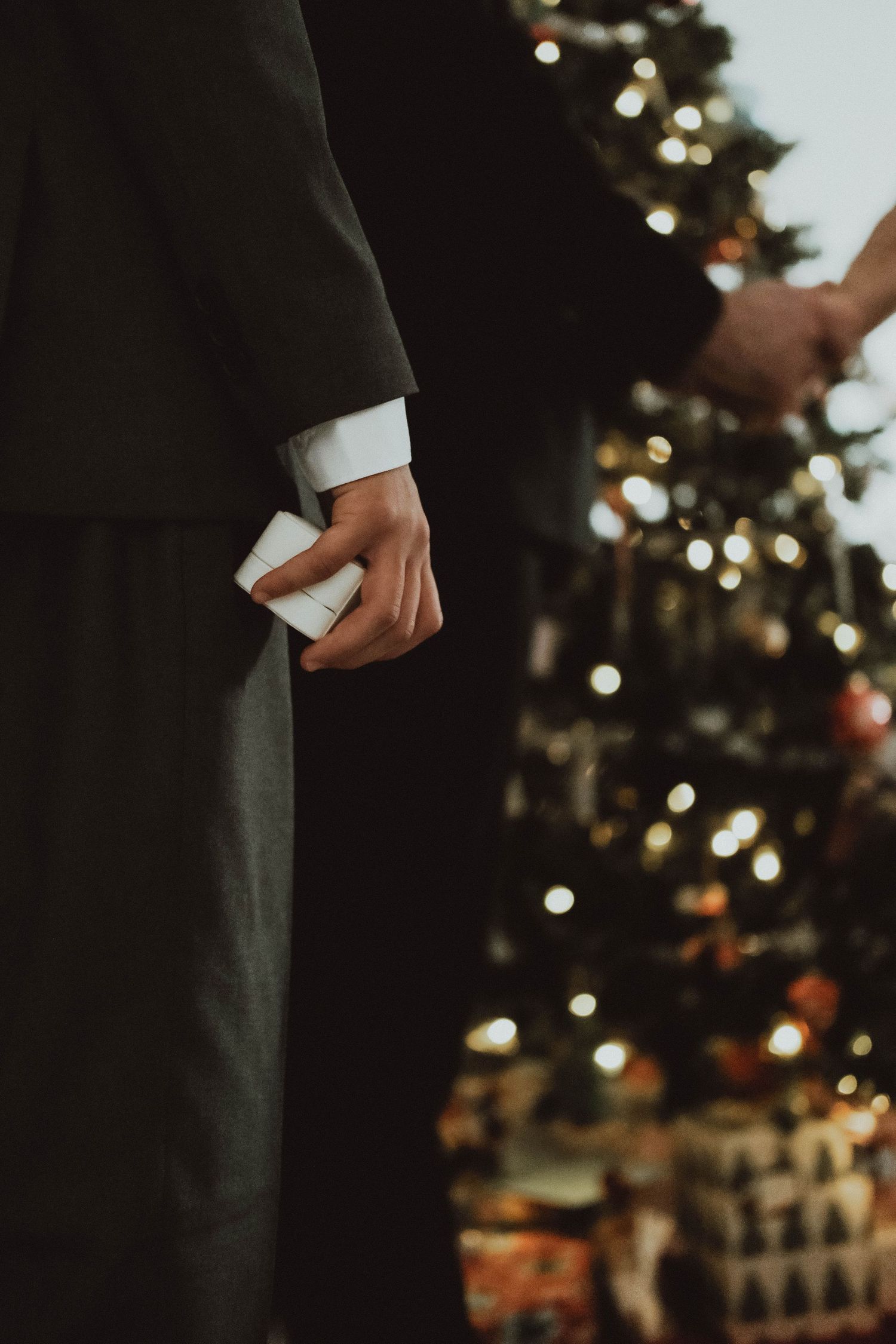 A hand holds a white ring box in front of a blurred Christmas tree with twinkling lights.