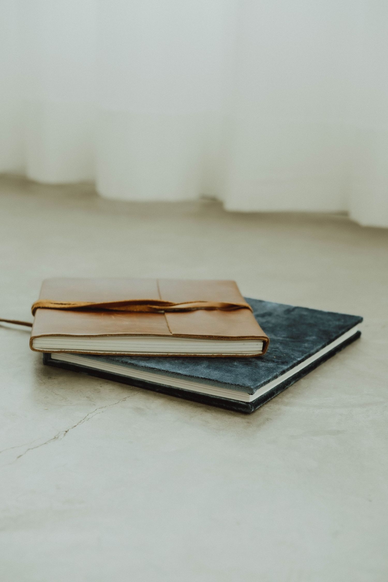 Two vintage notebooks with worn leather covers stacked on a white surface near a sheer curtain in soft lighting.