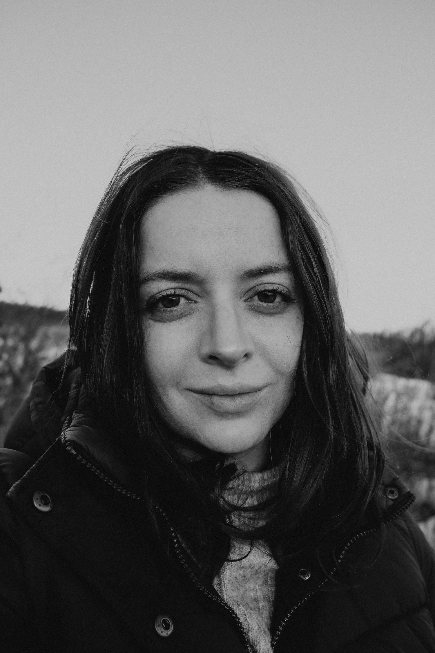 Black and white portrait of someone in a dark winter coat smiling at the camera outdoors.