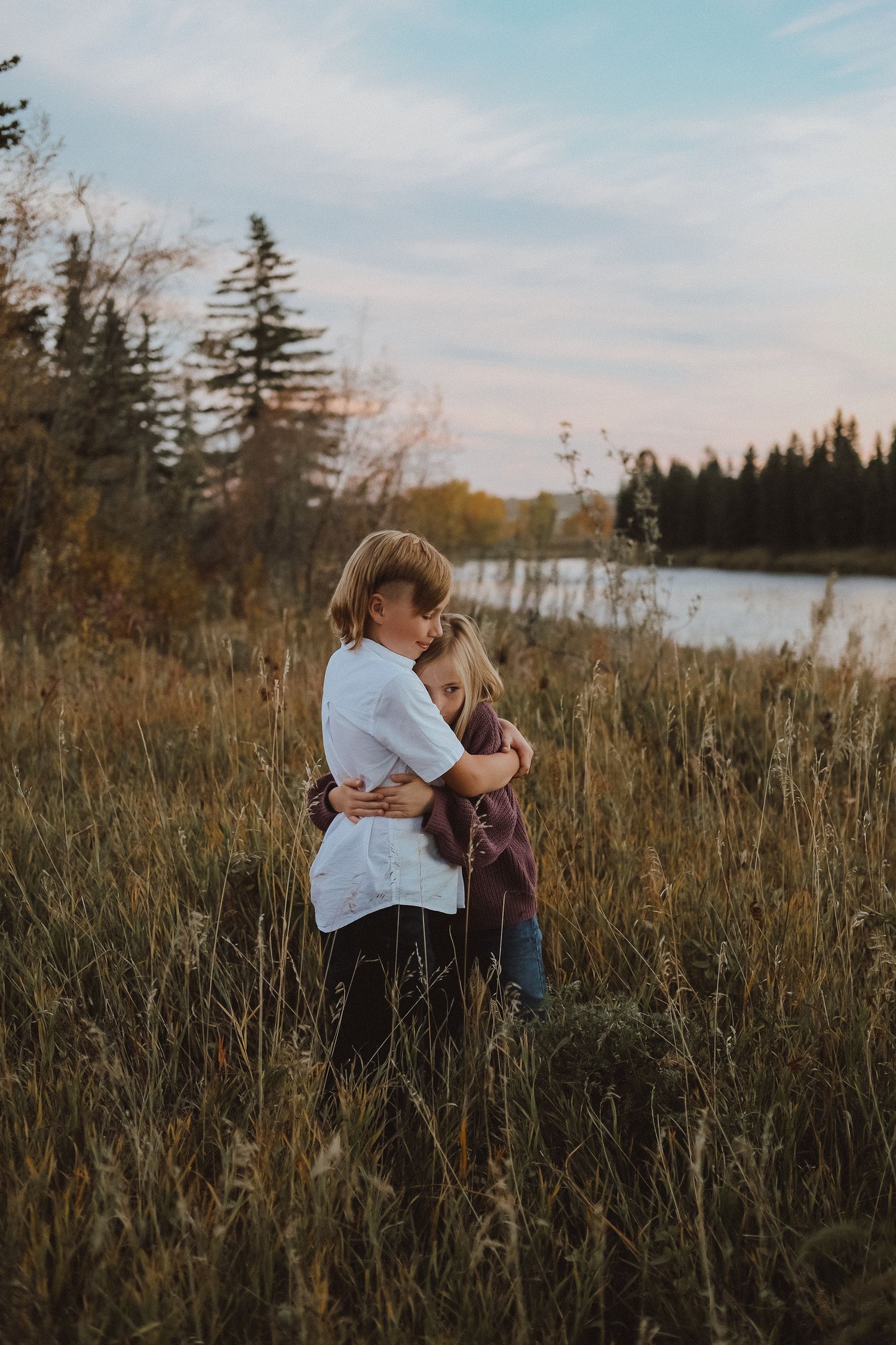 Two people embrace in a field of tall grass at sunset with evergreen trees and a lake in the background.