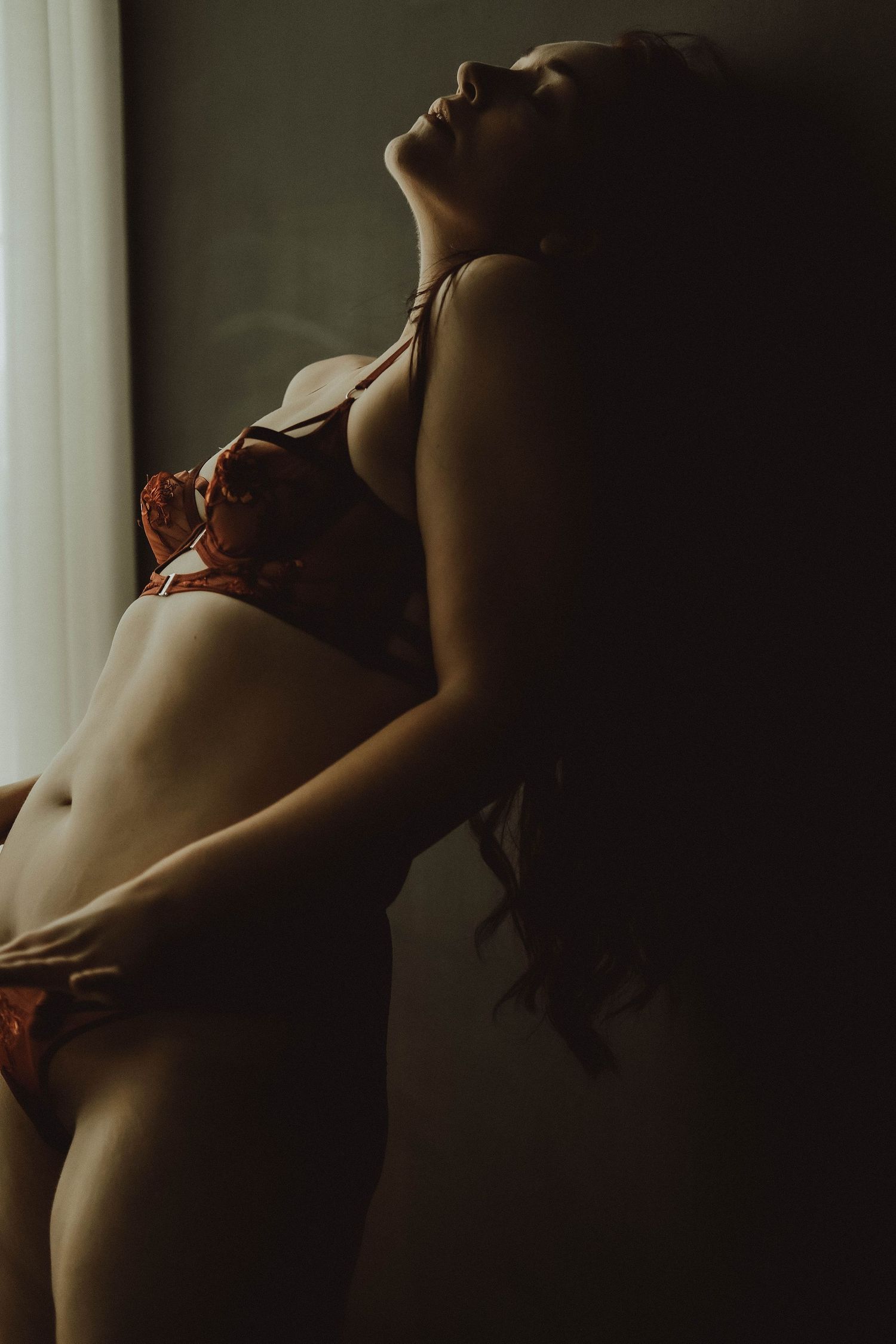 Artistic boudoir photograph with dramatic lighting shows a person in red lingerie posing against a window.