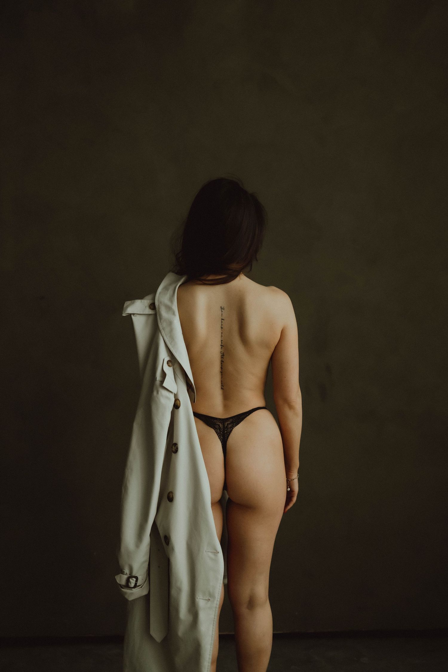 A nude figure stands against a dark wall wearing a white lab coat that hangs off their shoulders.