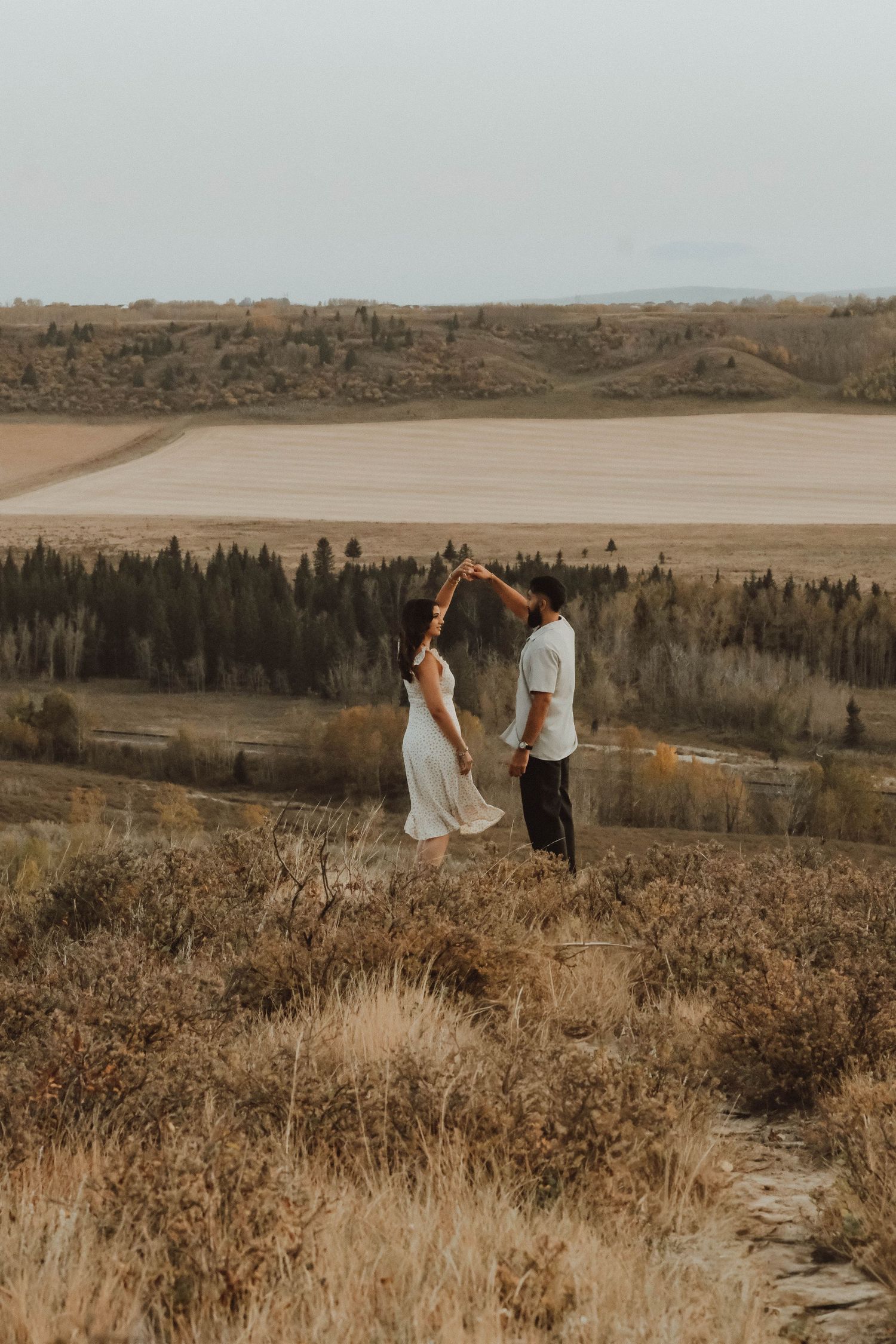 Two people stand together on a scenic hilltop overlooking a vast autumn landscape during golden hour.
