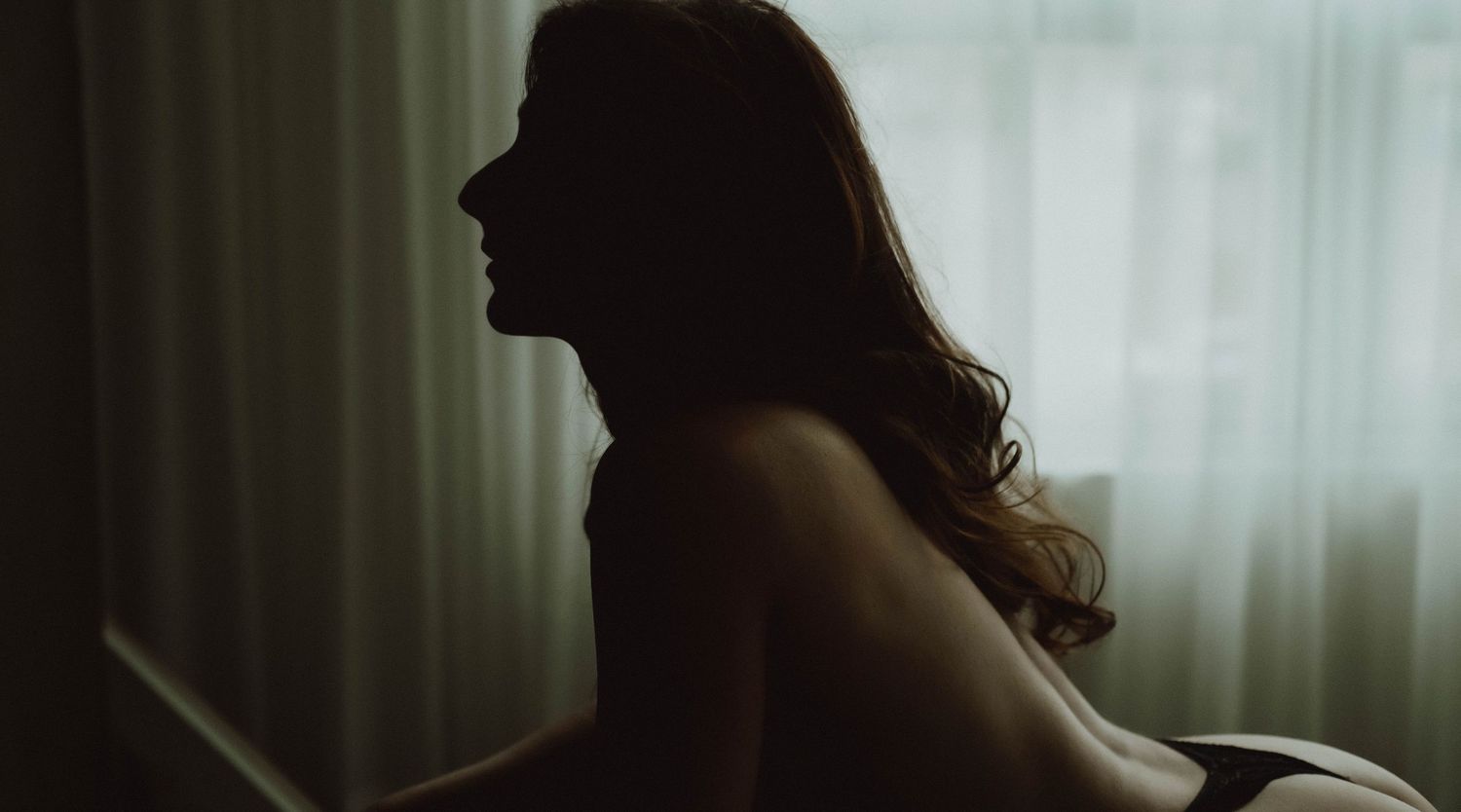 Silhouetted figure stands near window with dramatic backlighting creating a moody atmospheric scene.