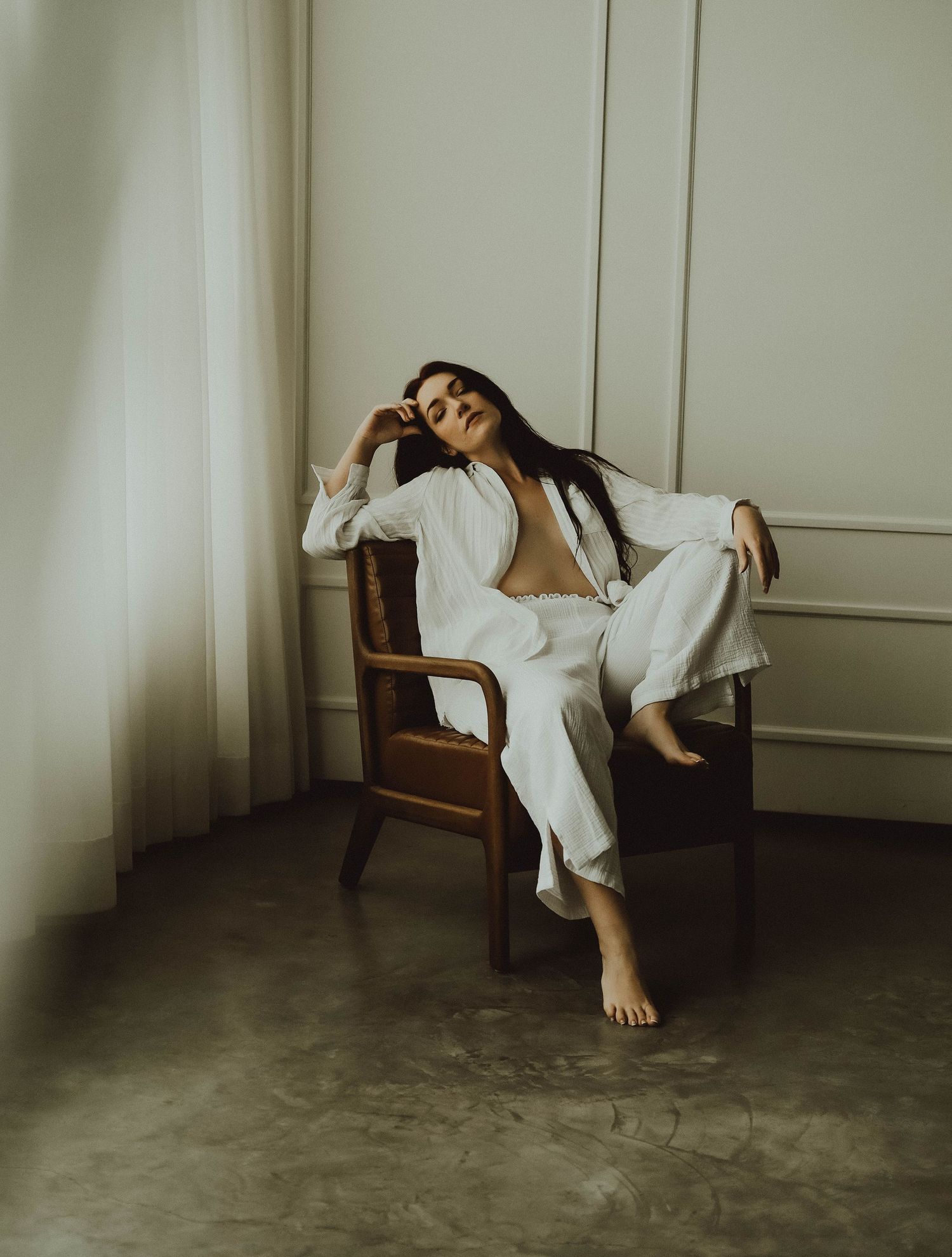 A figure in a white dress reclines dramatically across a wooden chair against a minimalist beige wall.