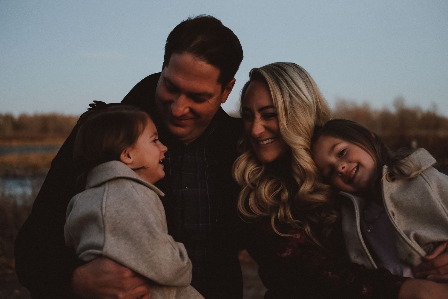 A family of four shares a warm embrace outdoors during golden hour, laughing and smiling together.