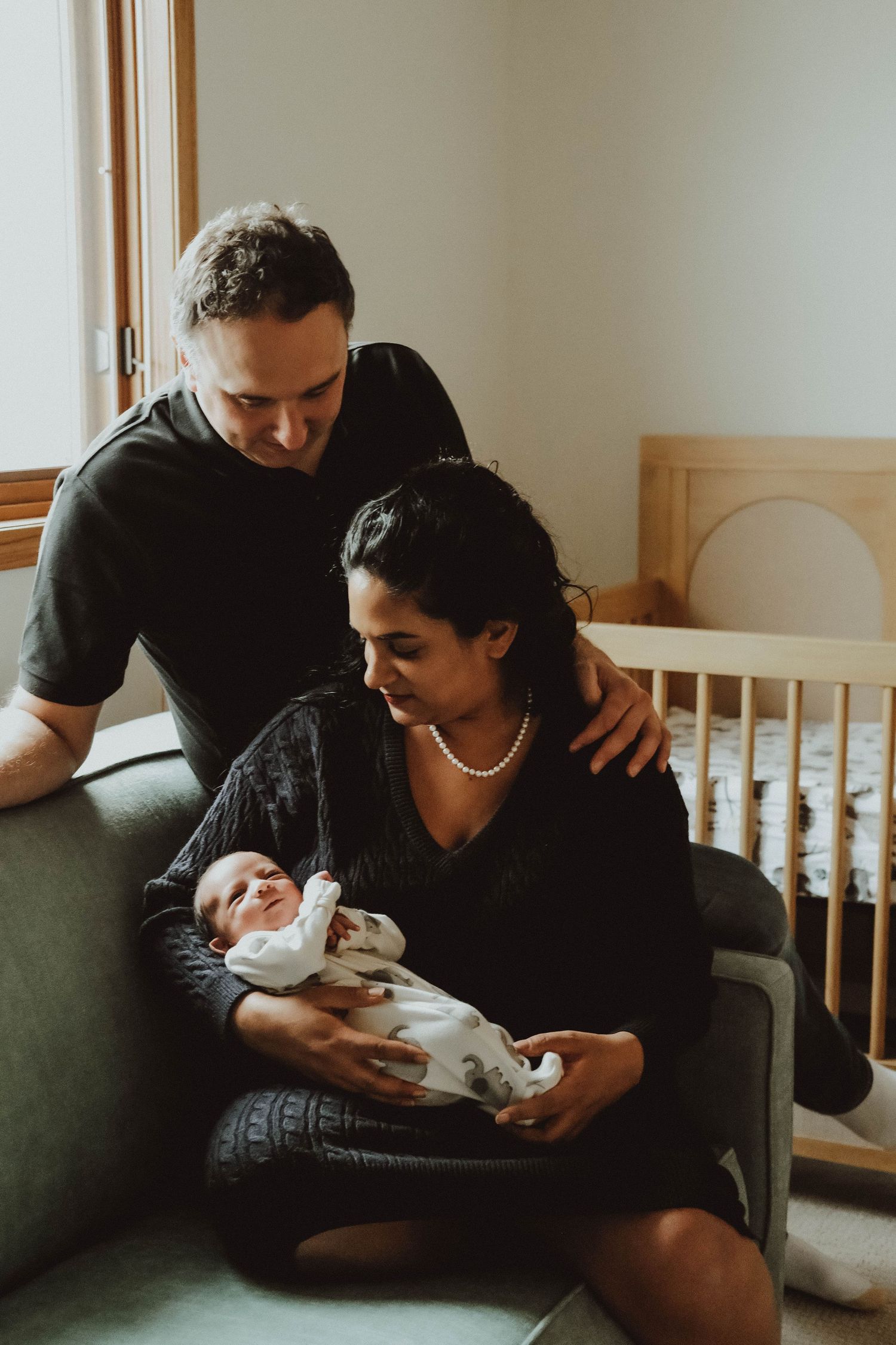 A couple sits together on a couch bonding with their newborn baby in a warm, natural light setting.