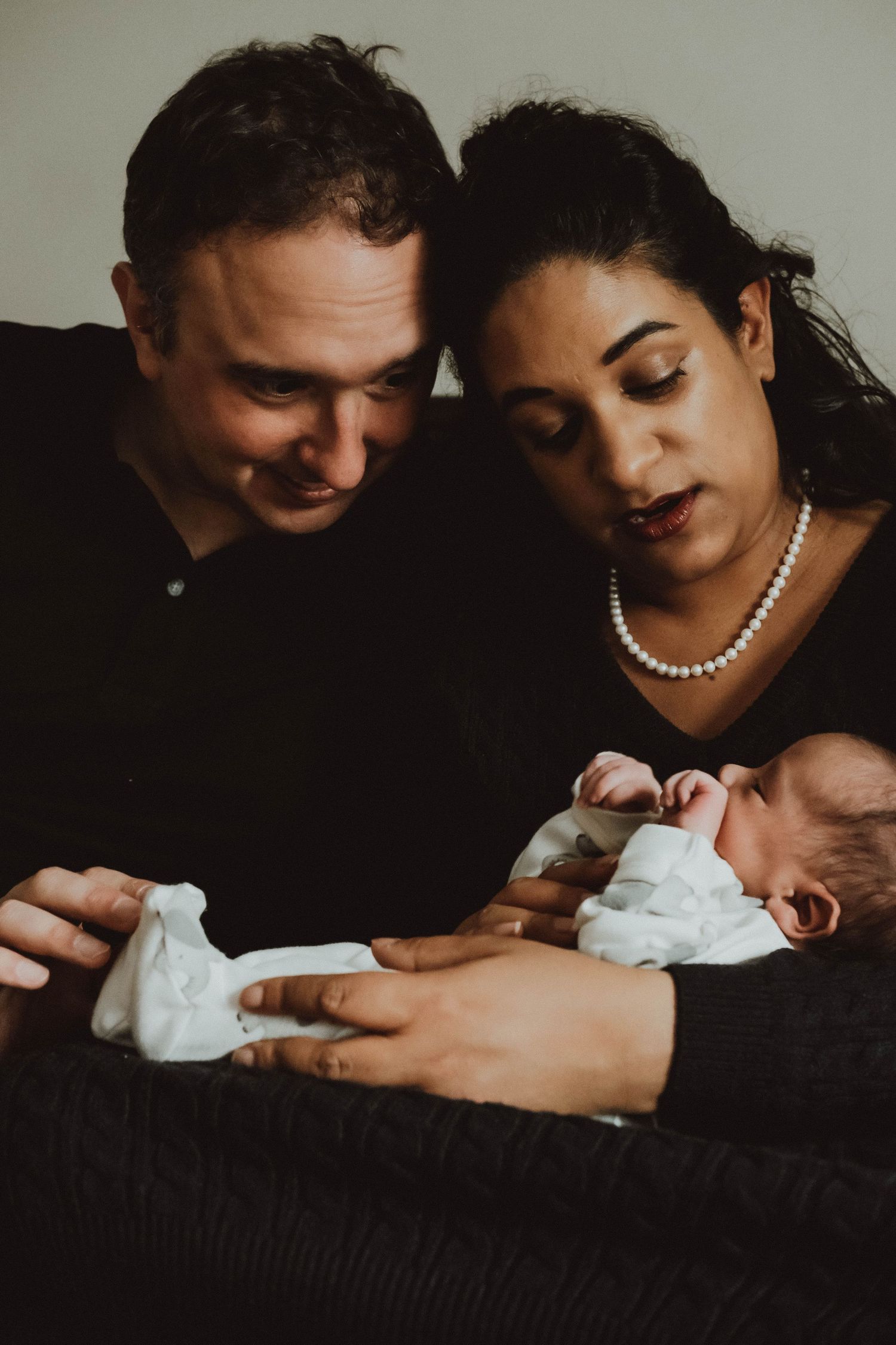 A couple shares an intimate moment with their newborn baby in a series of dark, moody portraits against black background.