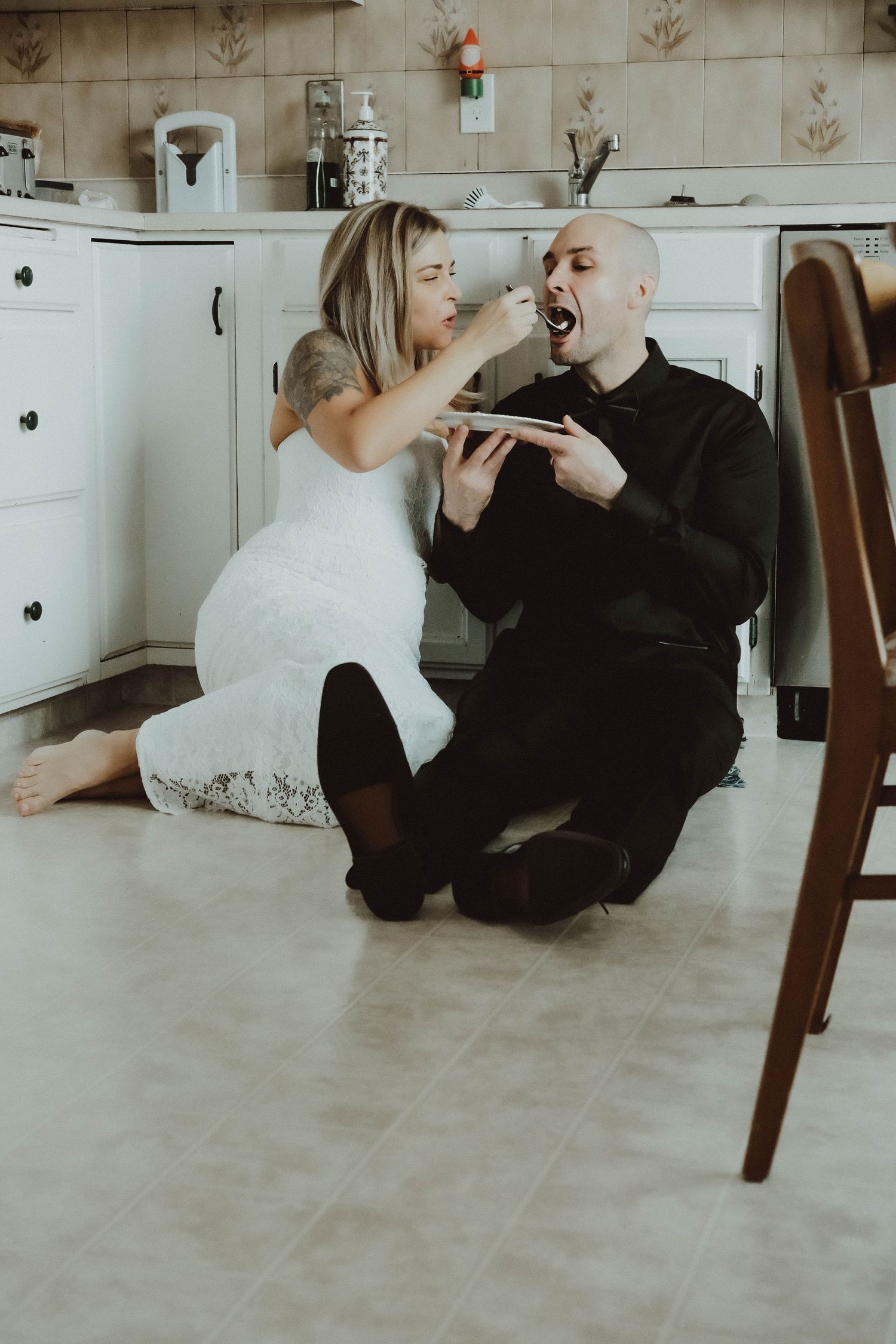 A bride in a white dress and groom in black share a playful moment sitting on the floor during their wedding shoot.