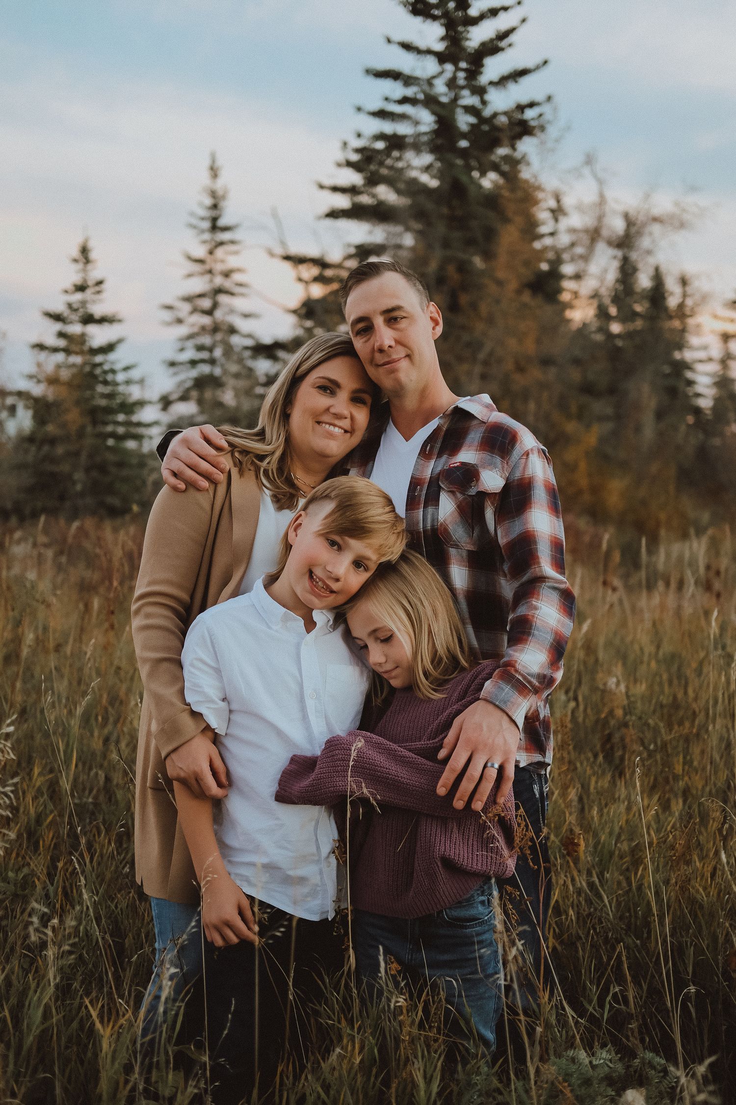 Family poses together in a field of tall grass with evergreen trees in the background during golden hour.