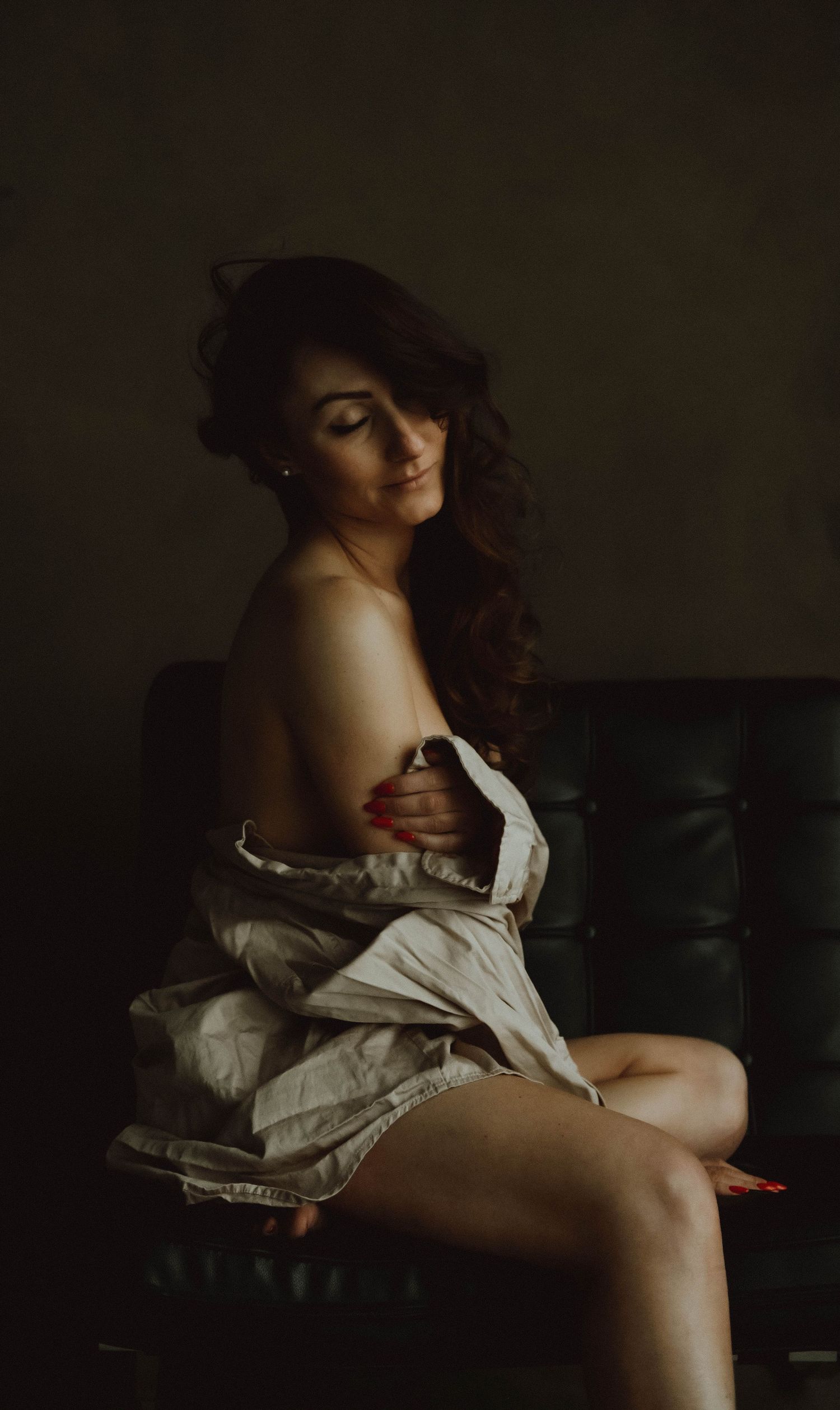 Moody boudoir portrait of woman in white dress sitting on dark leather chair with dramatic lighting.