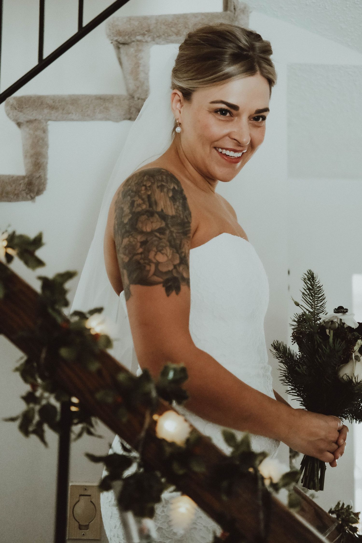 A bride in a white strapless wedding dress with a floral arm tattoo smiles while holding greenery by a staircase.