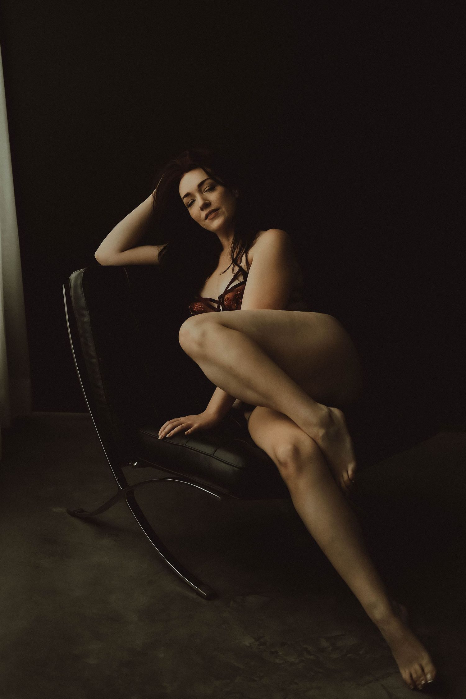 Artistic boudoir photo of woman in black lingerie reclining on modern leather chair in moody lighting.