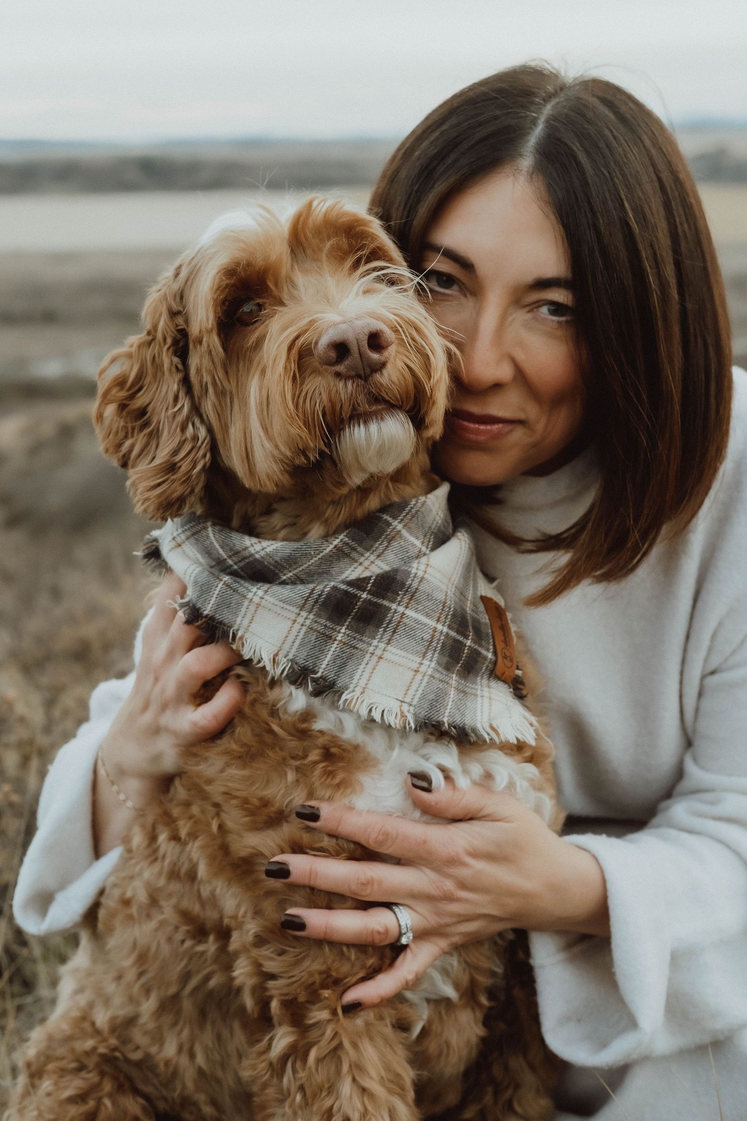 A brown Goldendoodle dog wearing a plaid bandana cuddles with someone in a cozy white sweater outdoors.