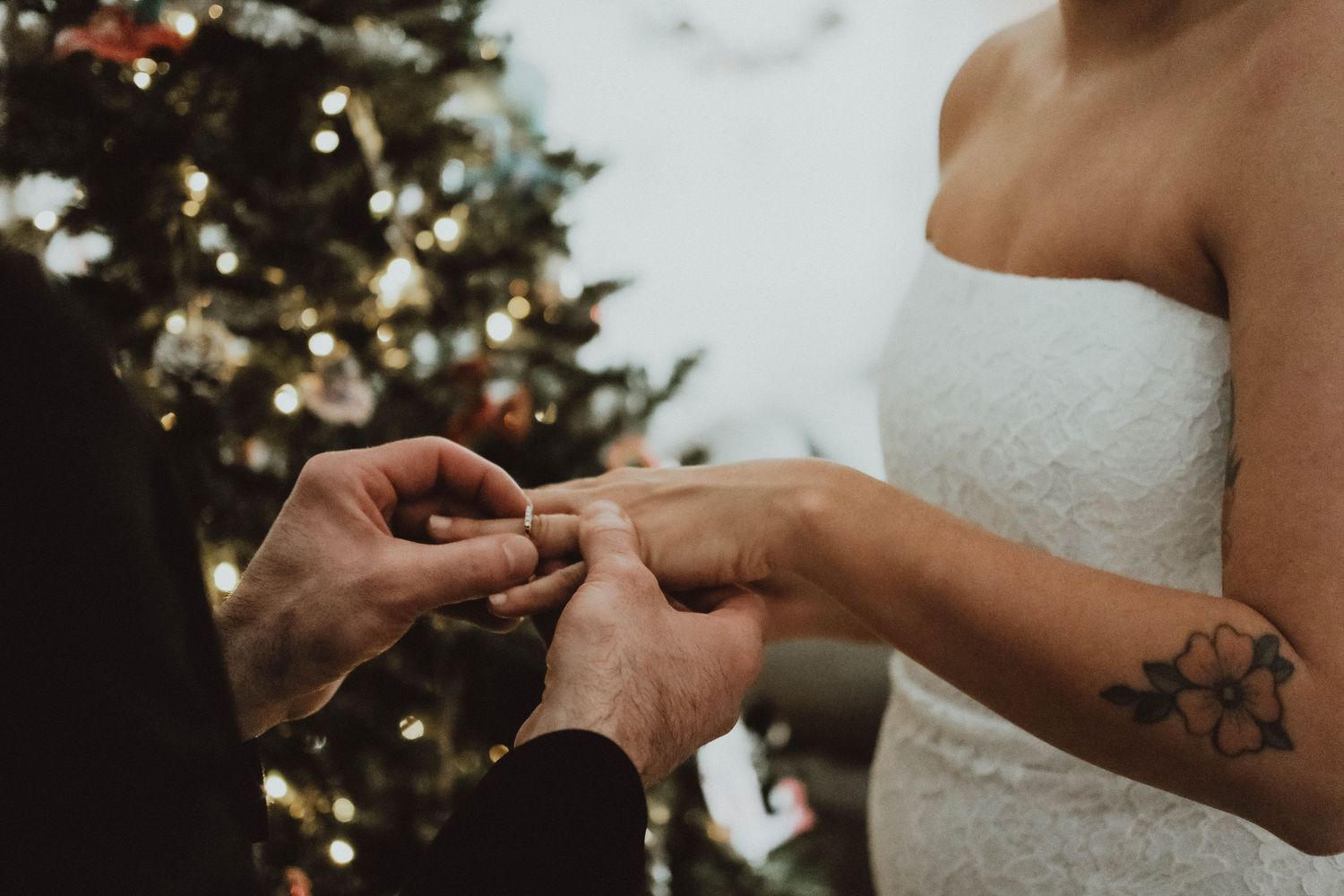 A close-up of hands exchanging wedding rings during a ceremony with a decorated Christmas tree in the background.