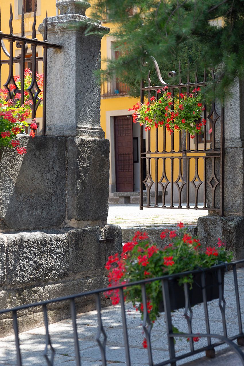 Stone gate pillars with red geranium flower baskets leading to a yellow Mediterranean building entrance.