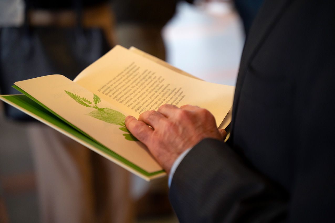 Man in suit reading open book with green illustrated pages at indoor event or ceremony.