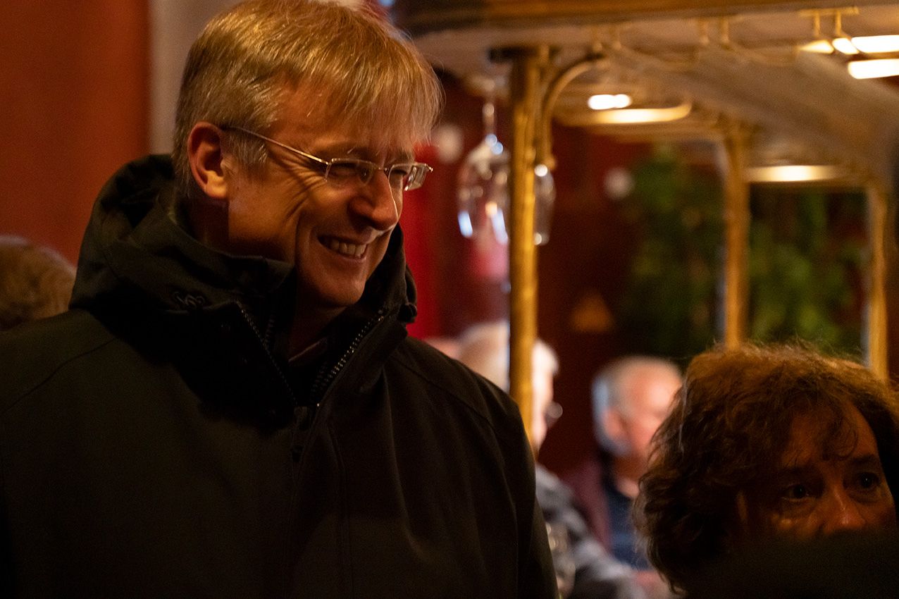 A smiling man wearing glasses enjoys a festive indoor event in a warmly lit decorated venue.