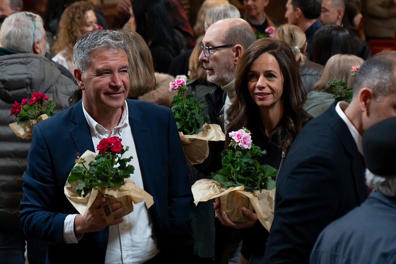 Smiling man and woman holding red and pink geranium bouquets wrapped in brown paper at indoor event.